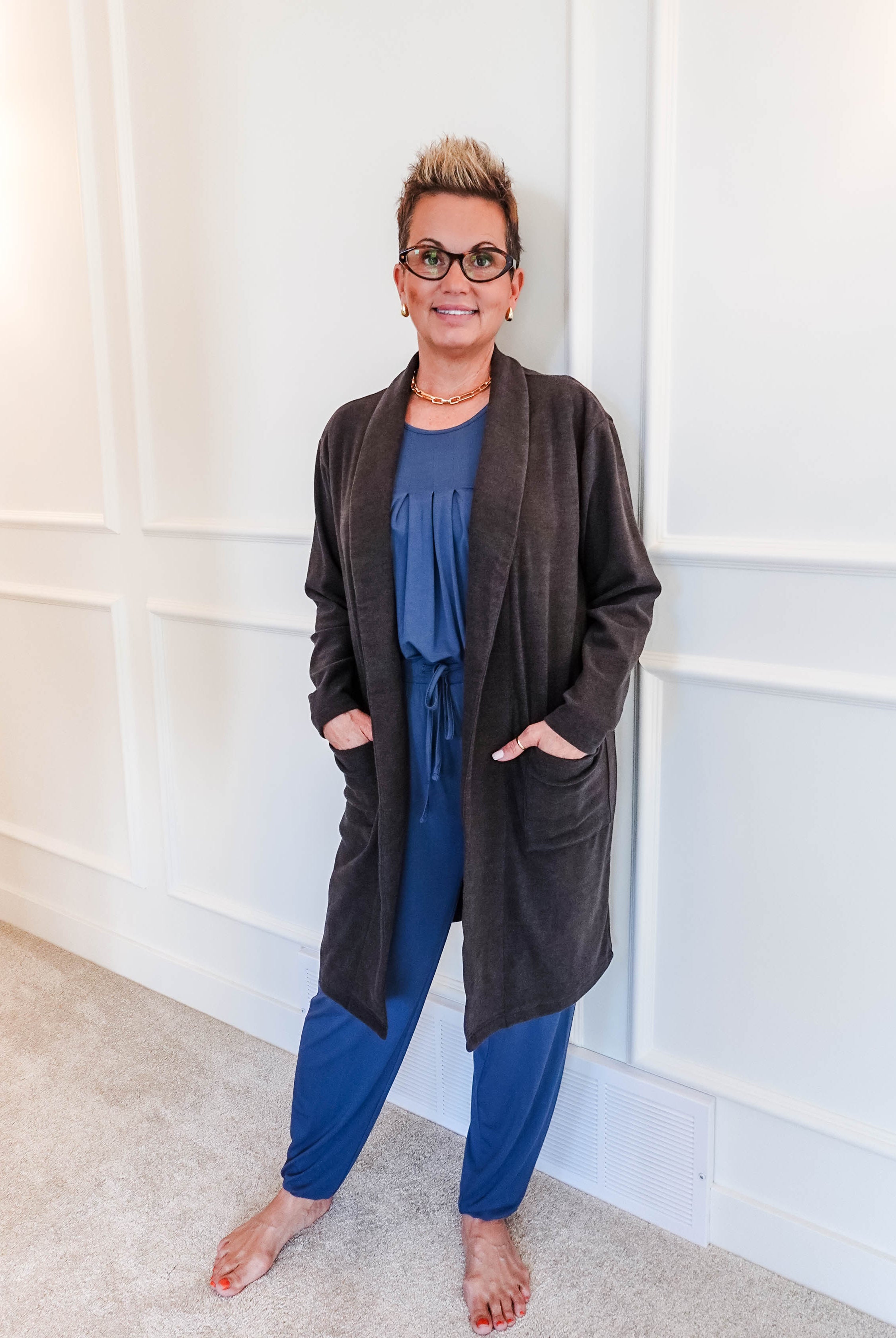 Full-length photo of a woman smiling, standing in a hallway, wearing a Charcoal Elevate Cardigan with deep patch pockets layered over a blue pyjama set. Classic collar, long-line structured fit.