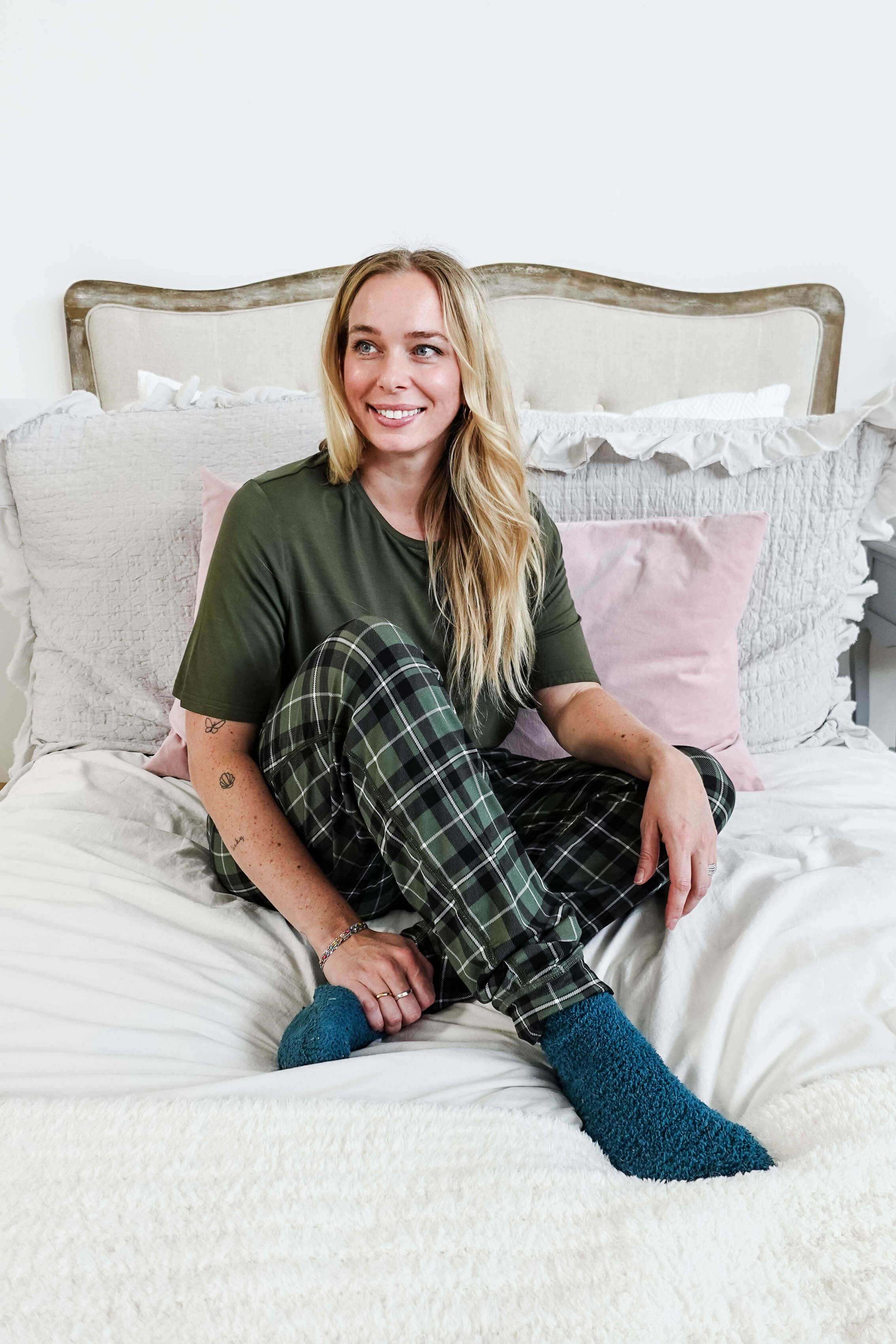 Woman sitting on a bed wearing Green Plaid Unisex Bamboo Jogger from Bob's Sleepwear Co. in a cozy bedroom setup.