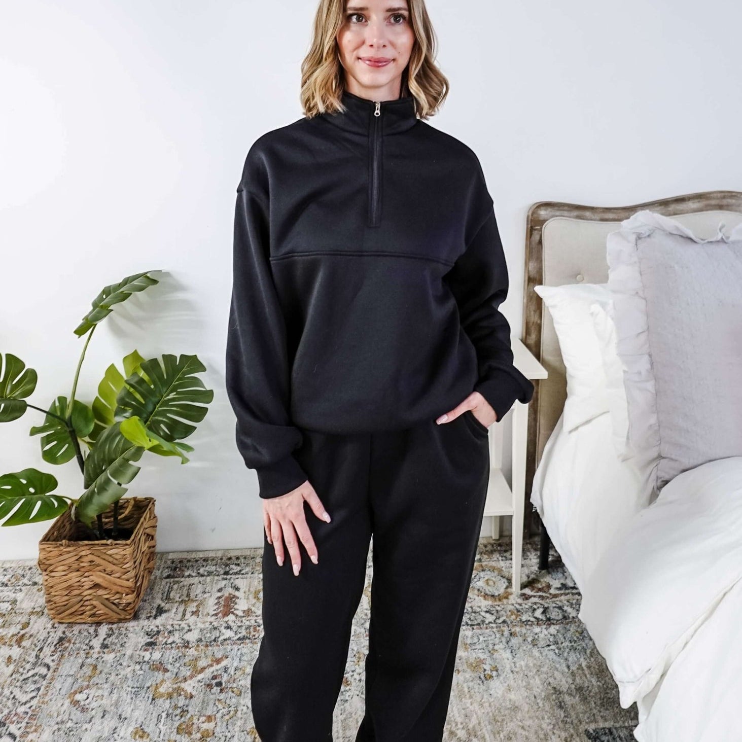 Woman wearing a black sweatsuit standing in a bedroom with a plant and bed in the background, Scarlett Ellie Half-Zip Sweatsuit Set | Cloud Nine Pajamas