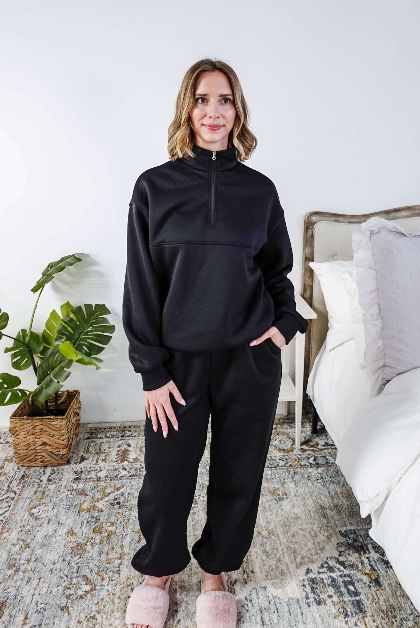 Woman wearing a black sweatsuit standing in a bedroom with a plant and bed in the background, Scarlett Ellie Half-Zip Sweatsuit Set | Cloud Nine Pajamas