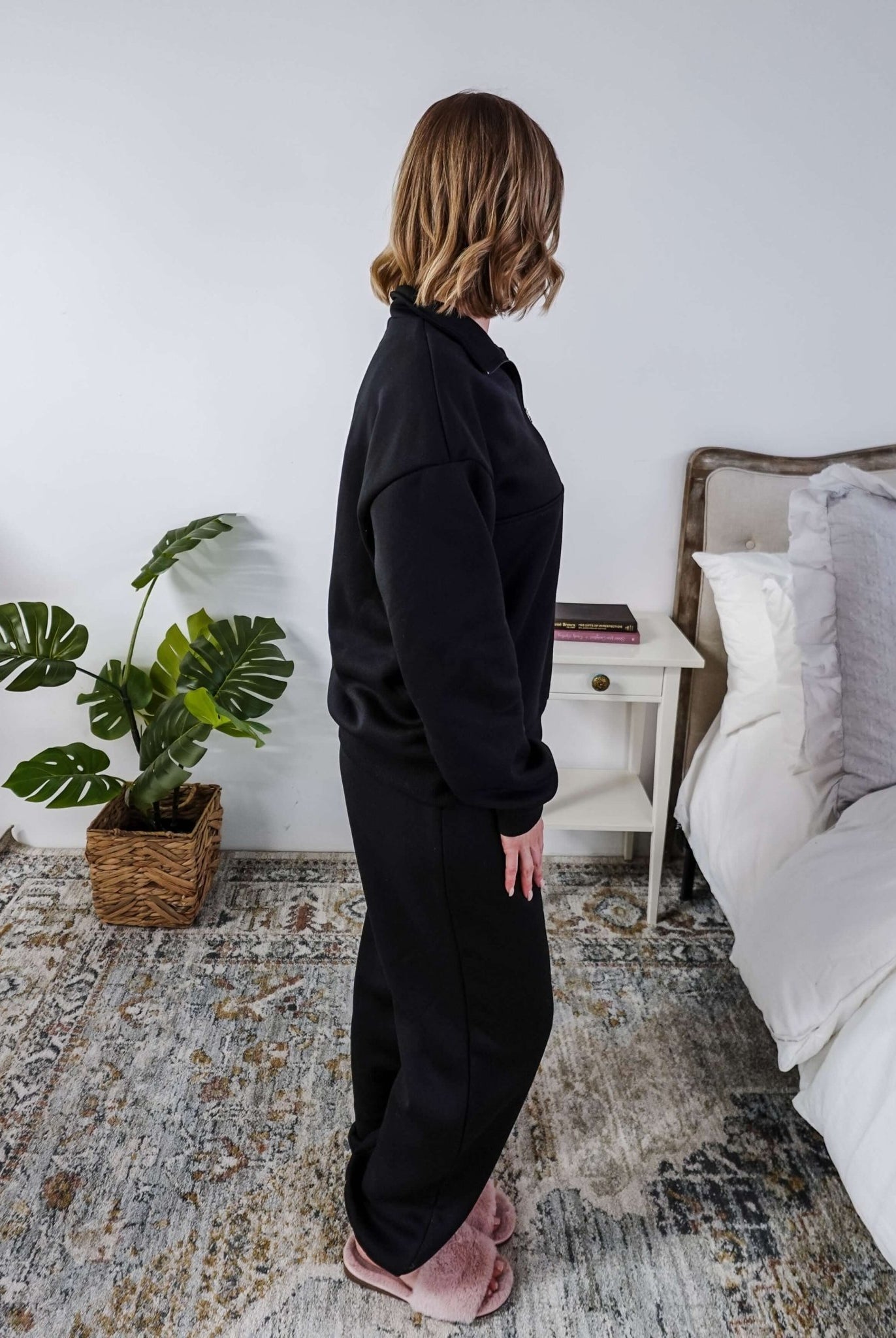 Woman wearing a black sweatsuit standing in a bedroom with a plant and bed in the background, Scarlett Ellie Half-Zip Sweatsuit Set | Cloud Nine Pajamas
