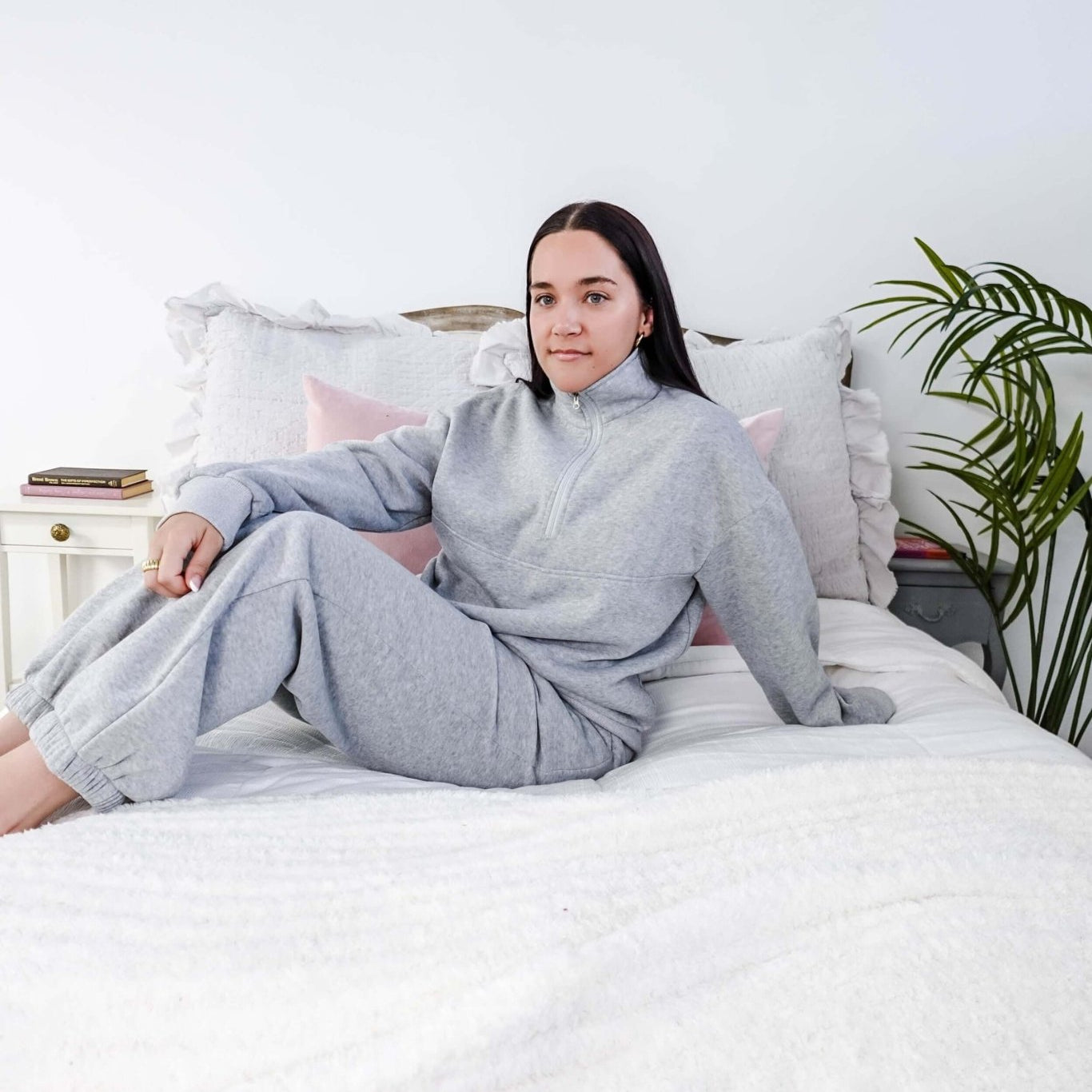 Person wearing a gray outfit sitting on a bed in a room with a plant. Scarlett Ellie Half-Zip Sweatsuit Set | Cloud Nine Pajamas