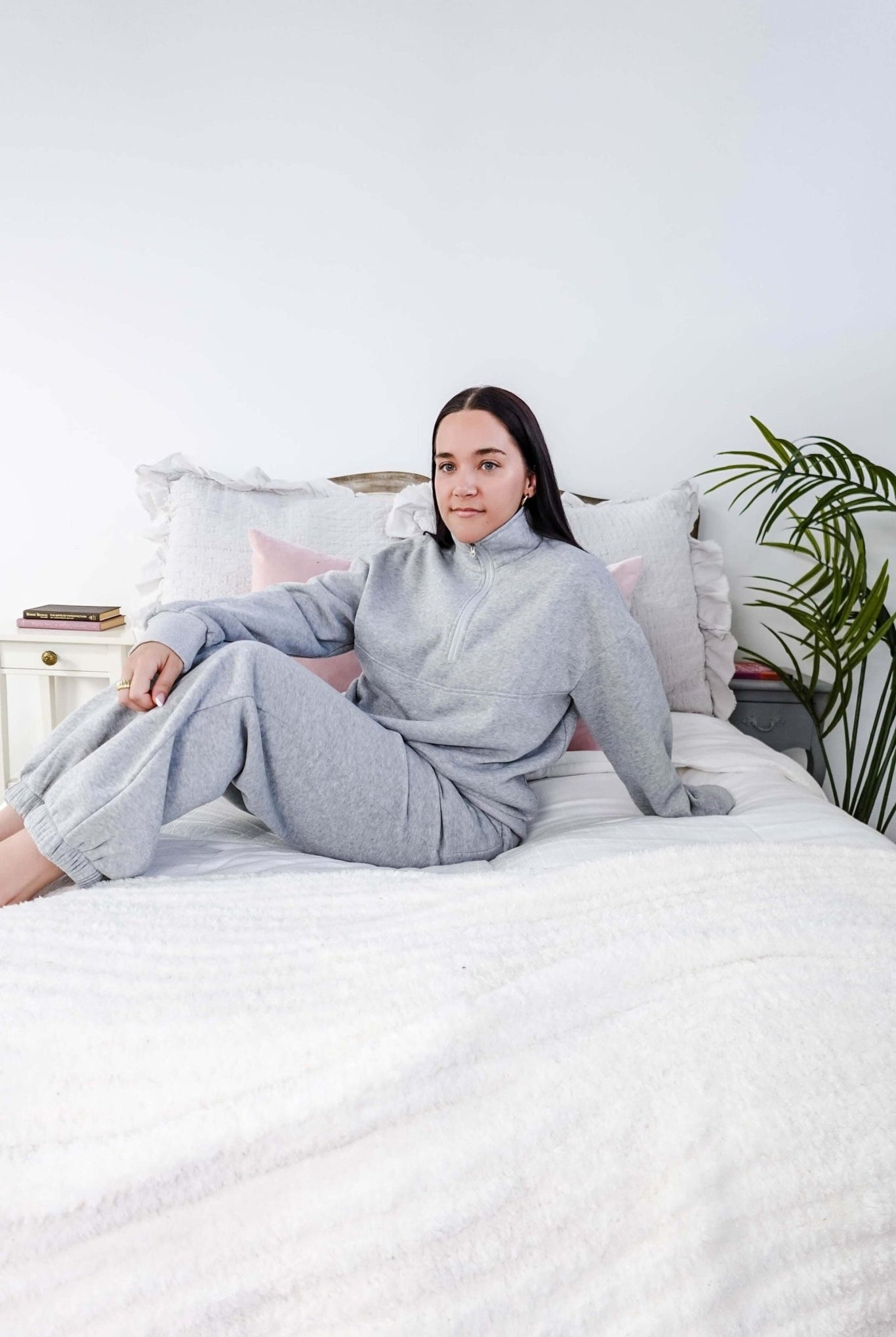 Person wearing a gray outfit sitting on a bed in a room with a plant. Scarlett Ellie Half-Zip Sweatsuit Set | Cloud Nine Pajamas