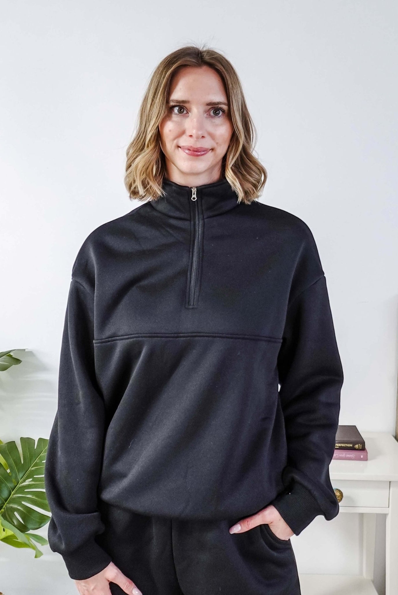 Woman wearing a black sweatsuit standing in a bedroom with a plant and bed in the background, Scarlett Ellie Half-Zip Sweatsuit Set | Cloud Nine Pajamas