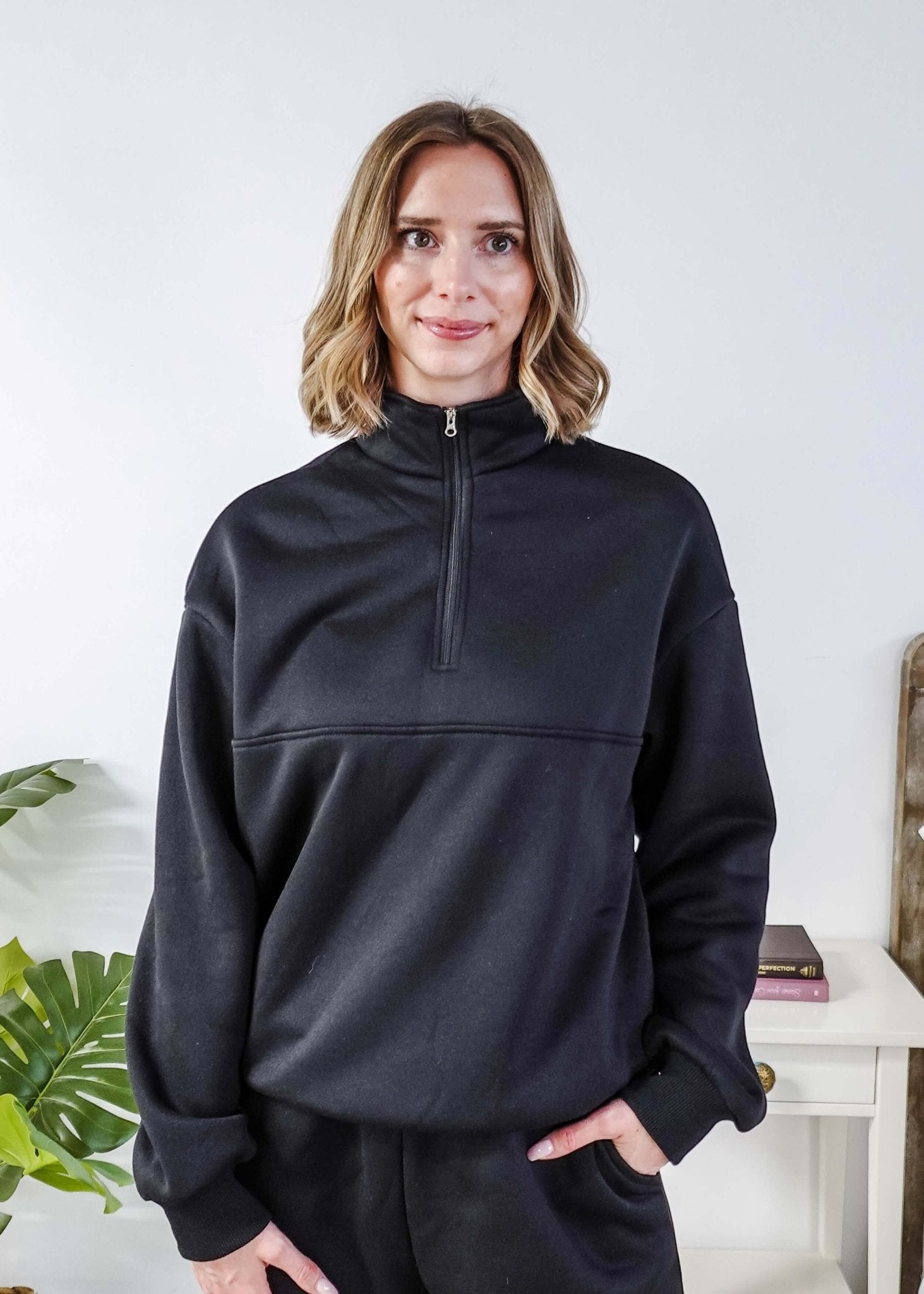 Woman wearing a black sweatsuit standing in a bedroom with a plant and bed in the background, Scarlett Ellie Half-Zip Sweatsuit Set | Cloud Nine Pajamas