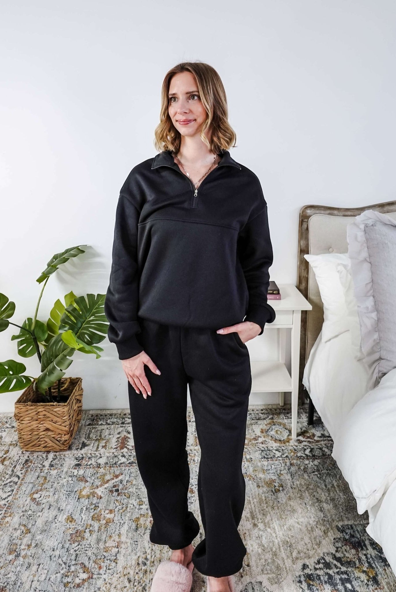 Woman wearing a black sweatsuit standing in a bedroom with a plant and bed in the background, Scarlett Ellie Half-Zip Sweatsuit Set | Cloud Nine Pajamas