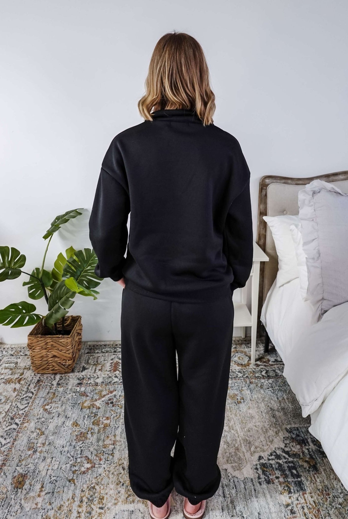 Woman wearing a black sweatsuit standing in a bedroom with a plant and bed in the background, Scarlett Ellie Half-Zip Sweatsuit Set | Cloud Nine Pajamas