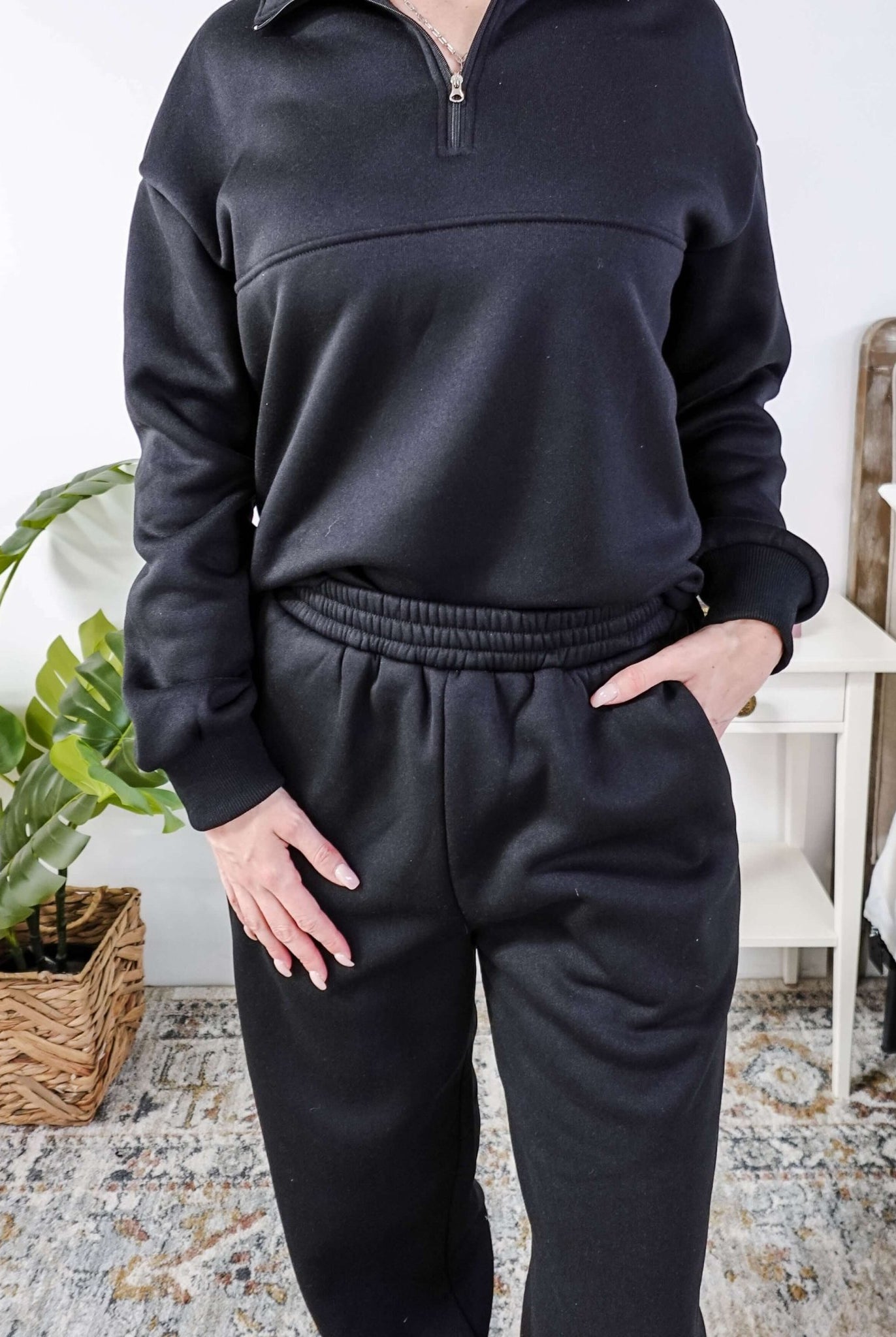 Woman wearing a black sweatsuit standing in a bedroom with a plant and bed in the background, Scarlett Ellie Half-Zip Sweatsuit Set | Cloud Nine Pajamas