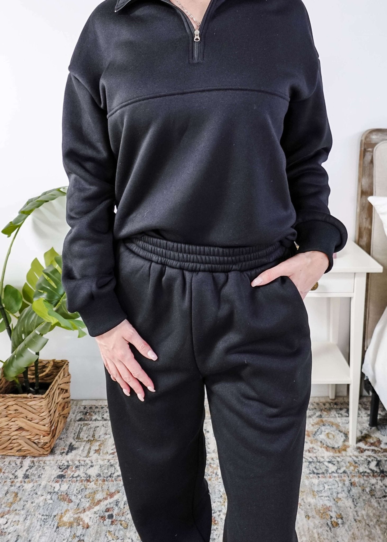 Woman wearing a black sweatsuit standing in a bedroom with a plant and bed in the background, Scarlett Ellie Half-Zip Sweatsuit Set | Cloud Nine Pajamas