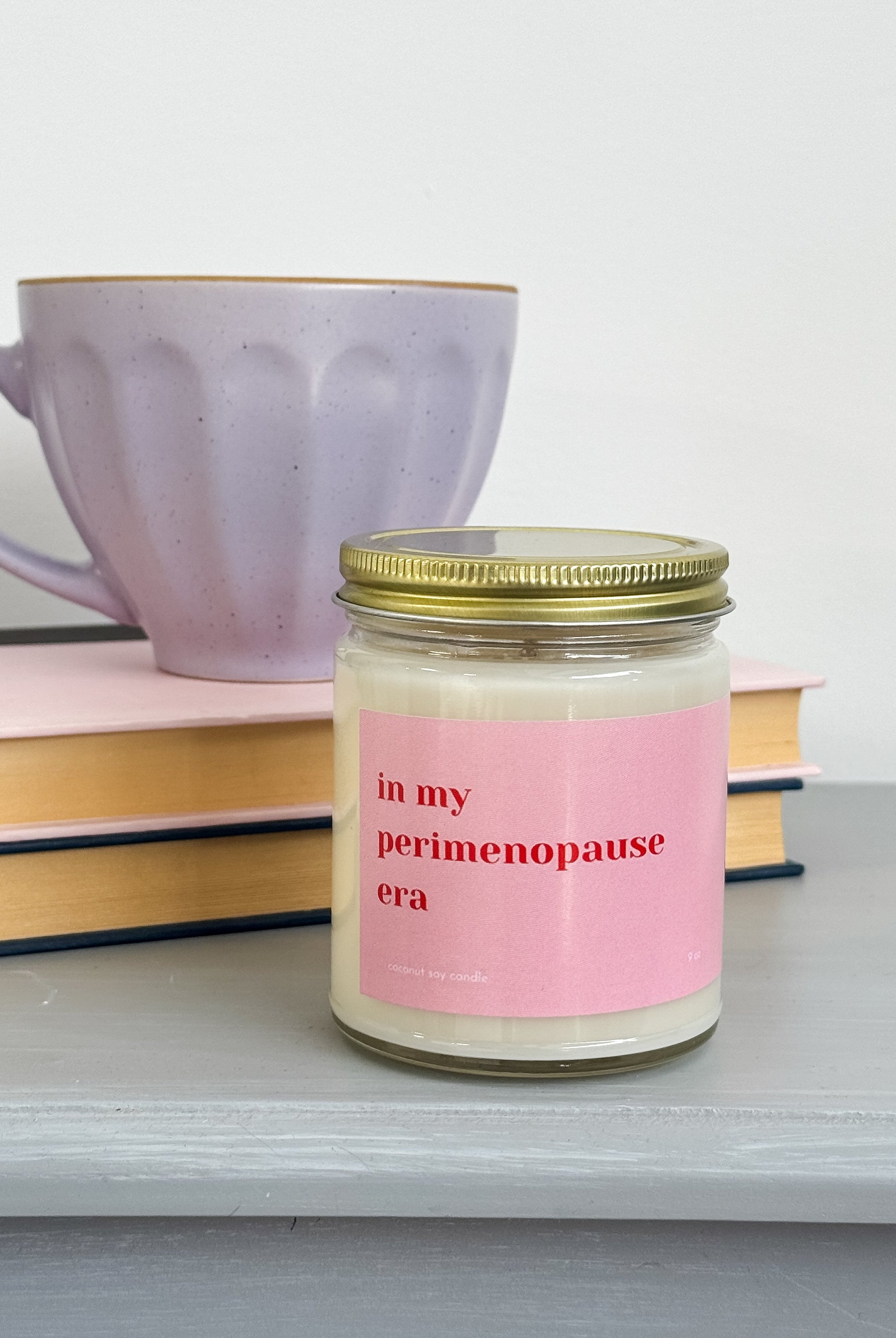 Jar of candle with 'in my perimenopause era' label on a surface with books and a mug by Heslip Home