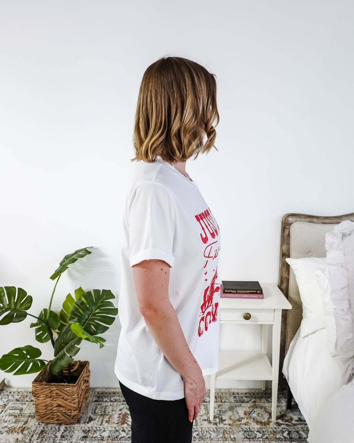 Woman wearing a white t-shirt with red text saying Just here for the cowboys in a bedroom setting