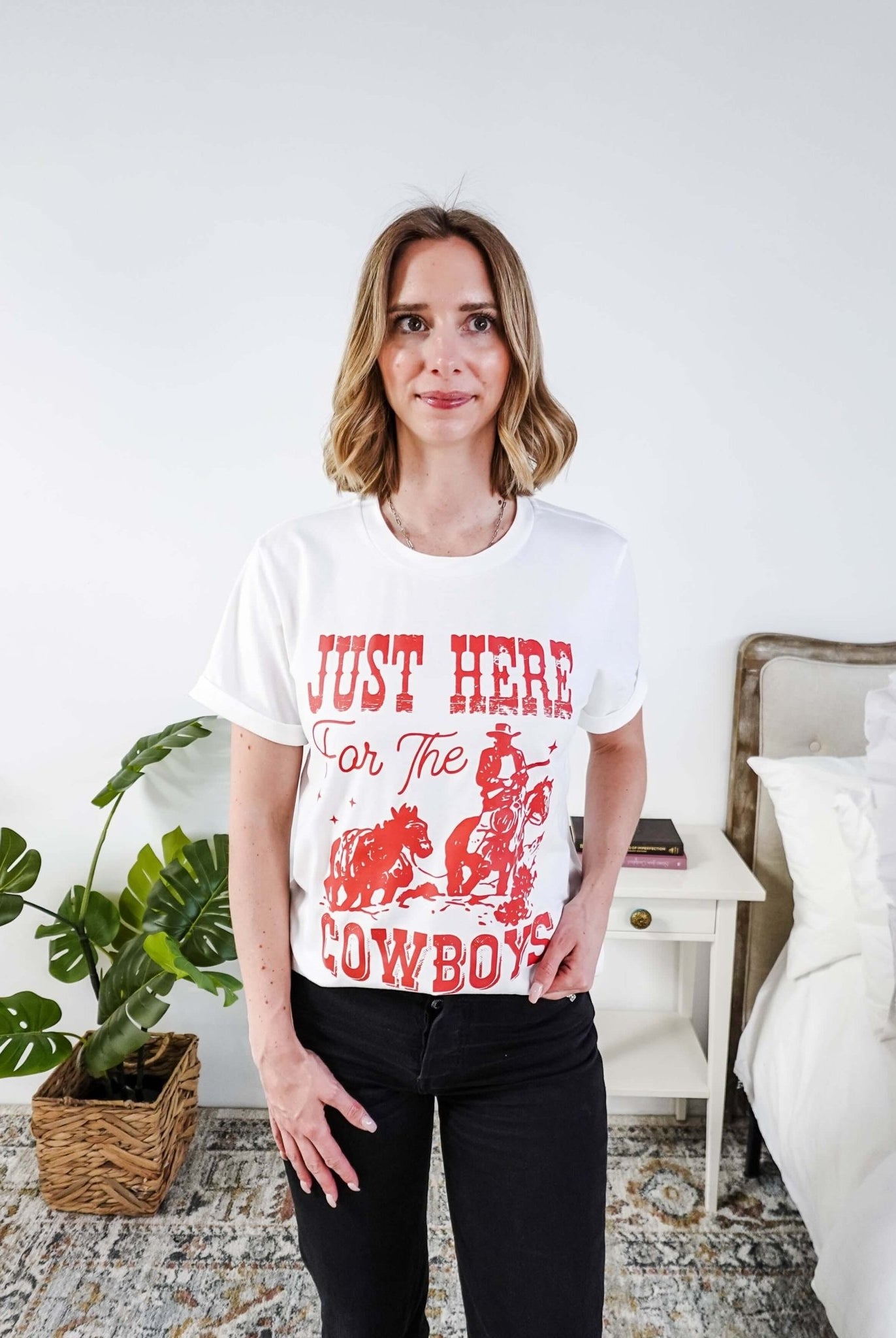 Woman wearing a white t-shirt with red text saying Just here for the cowboys in a bedroom setting
