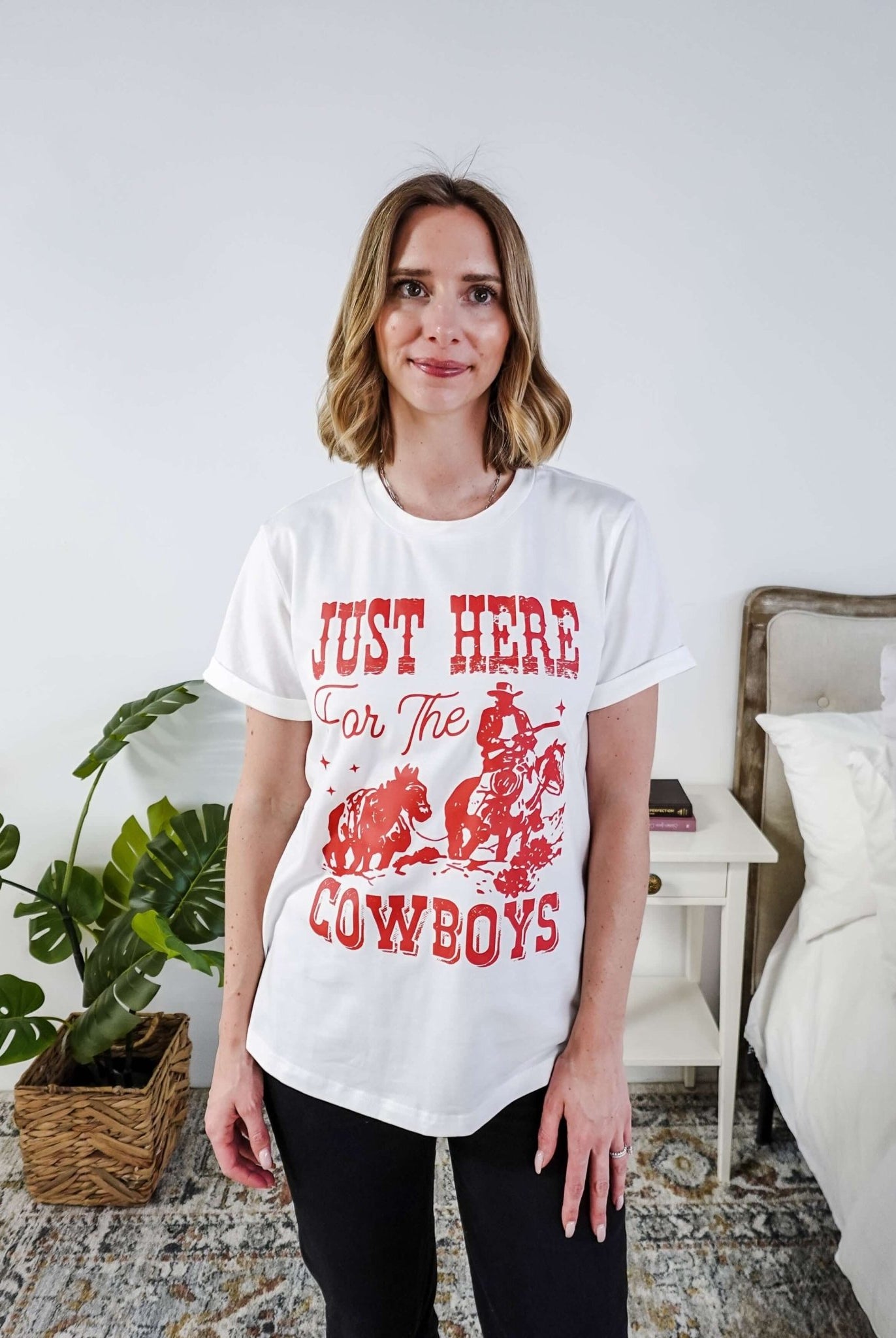 Woman wearing a white t-shirt with red text saying Just here for the cowboys in a bedroom setting
