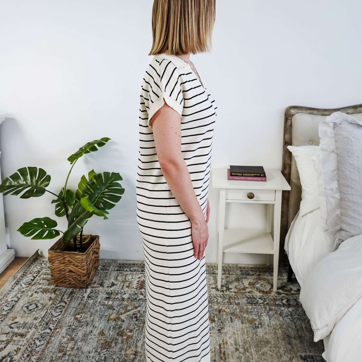 Woman in a striped black and white knit dress standing in a bedroom with a plant and bed in the background