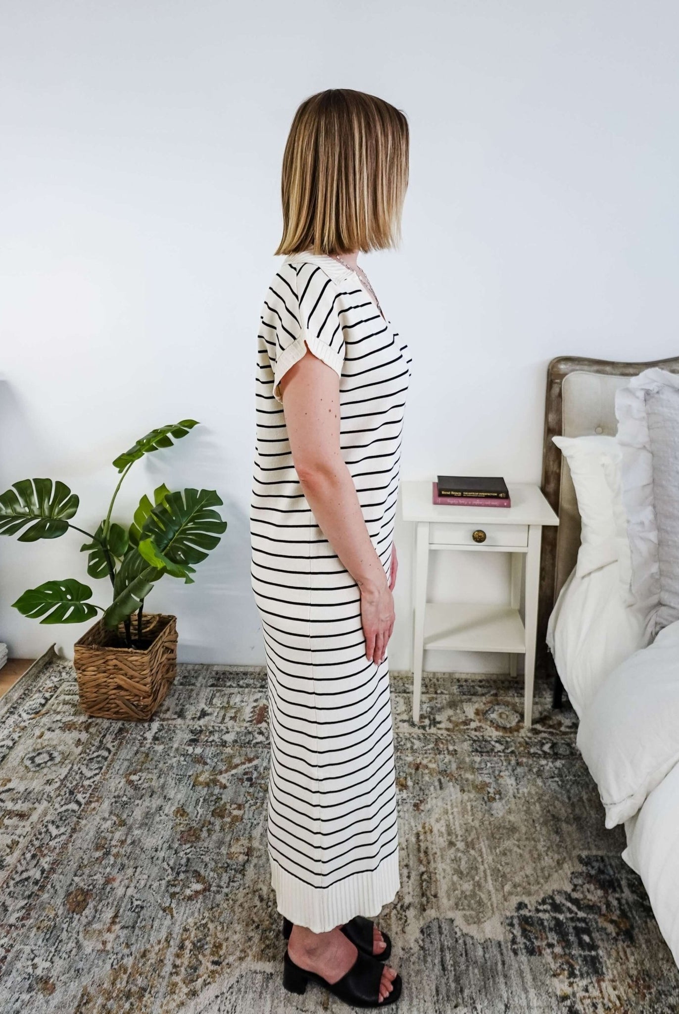 Woman in a striped black and white knit dress standing in a bedroom with a plant and bed in the background