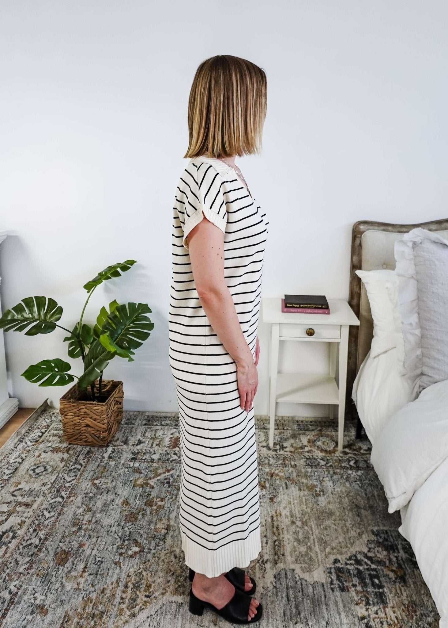 Woman in a striped black and white knit dress standing in a bedroom with a plant and bed in the background