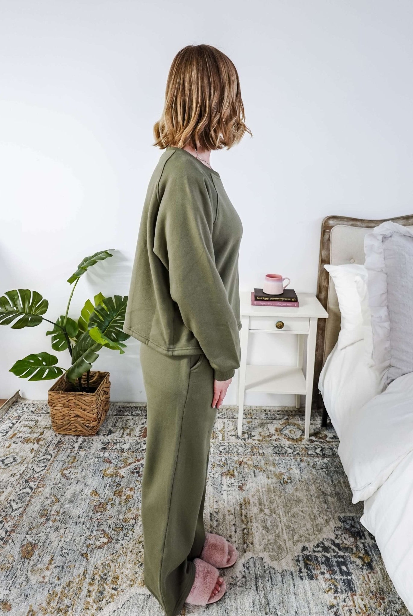 Woman in green pajama set standing in a bedroom with a plant and nightstand.