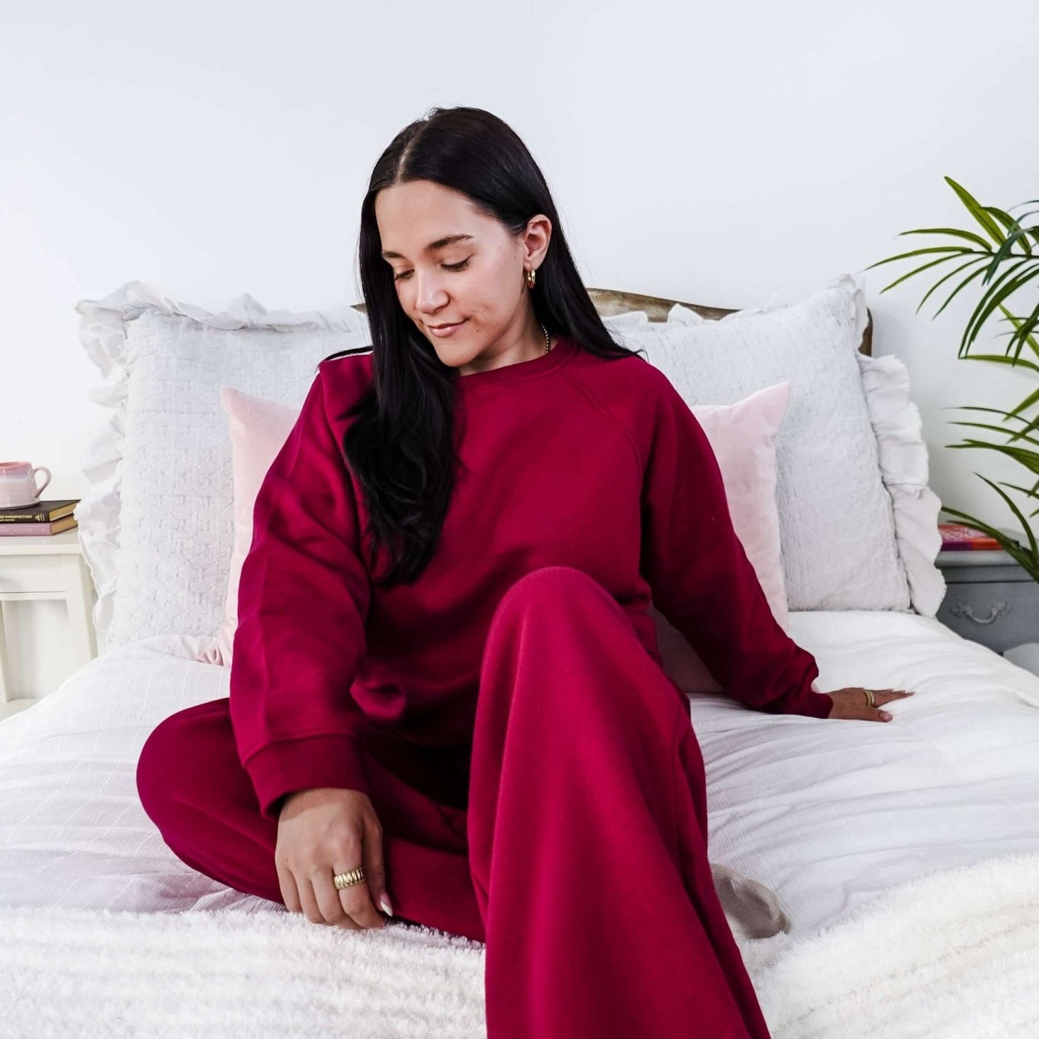 Woman in a red sweatsuit sitting on a bed in a bright room with white walls and a plant.
