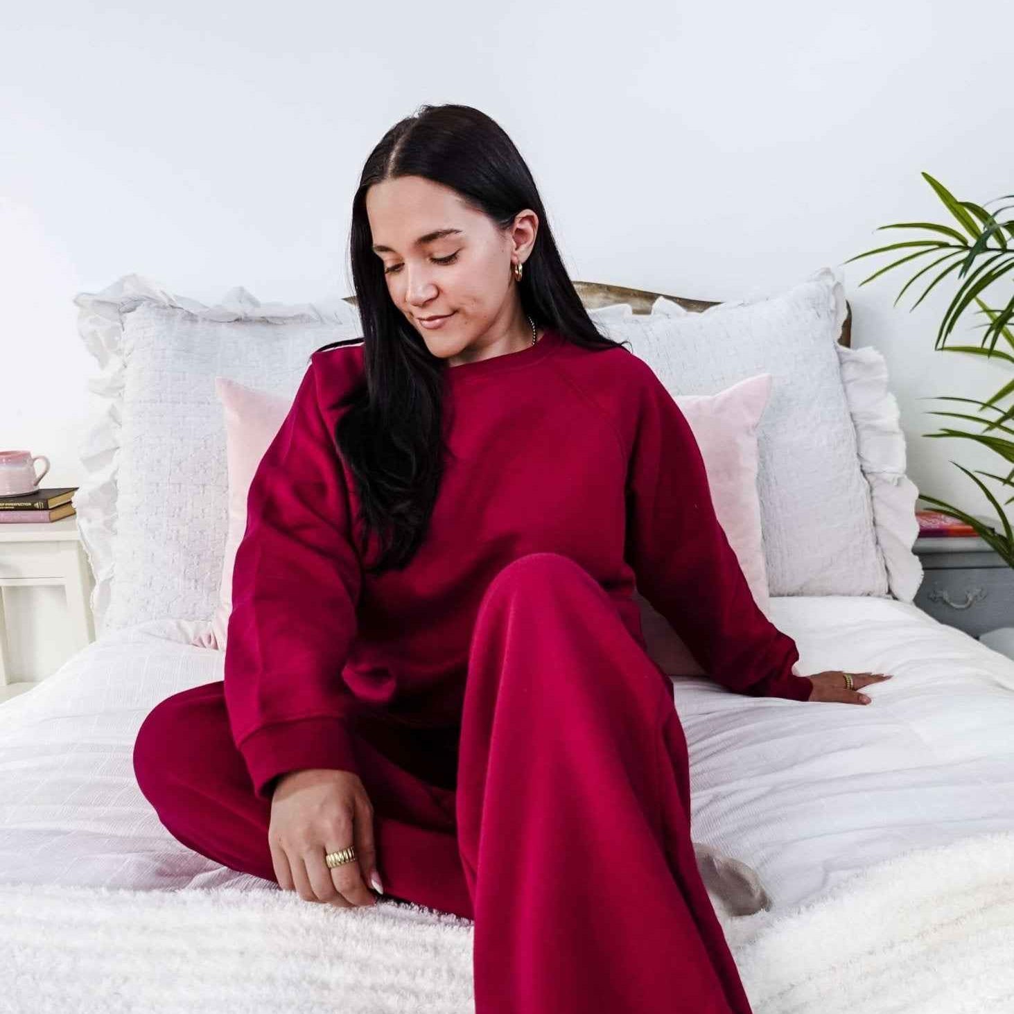 Woman in a red sweatsuit sitting on a bed in a bright room with white walls and a plant.