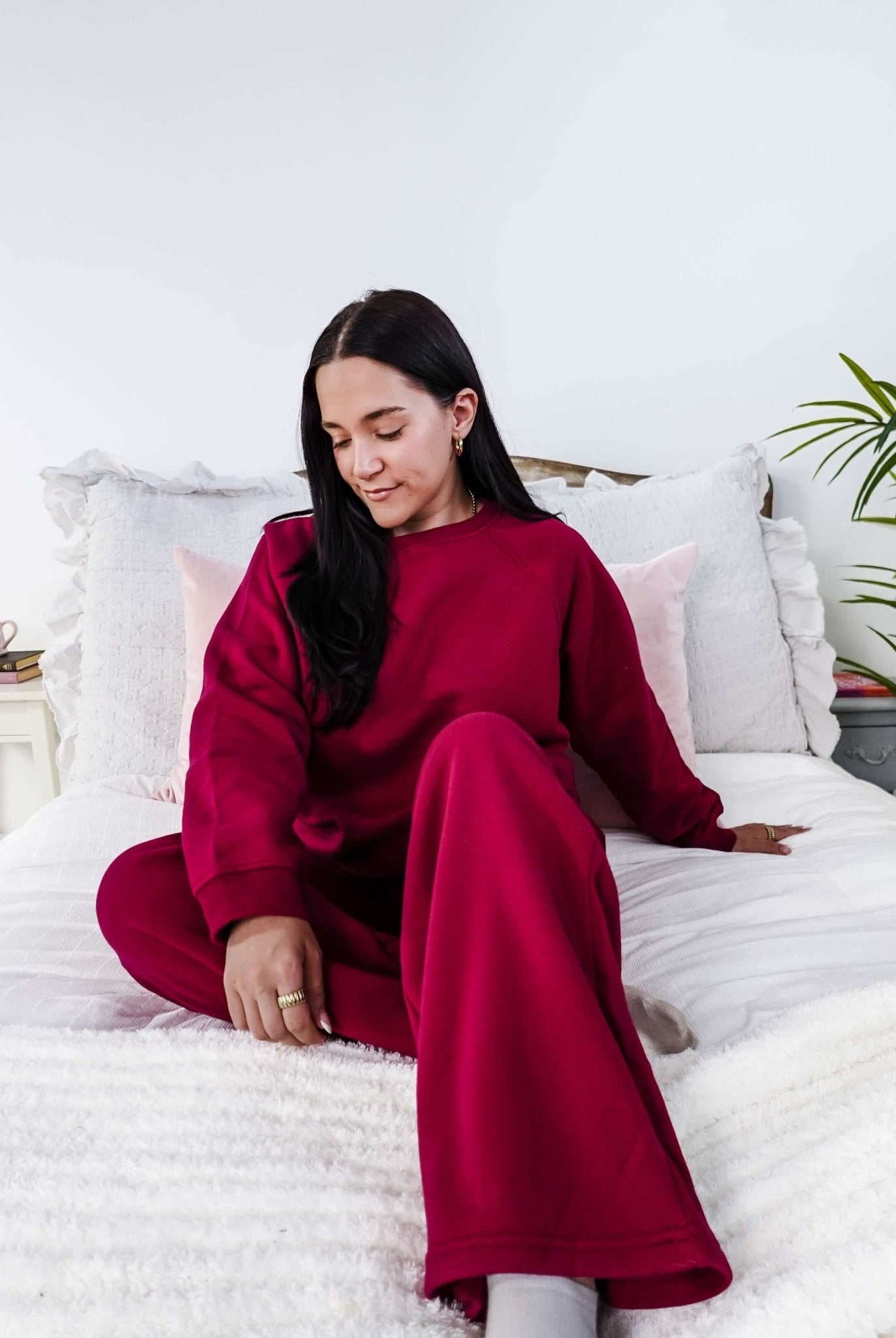 Woman in a red sweatsuit sitting on a bed in a bright room with white walls and a plant.