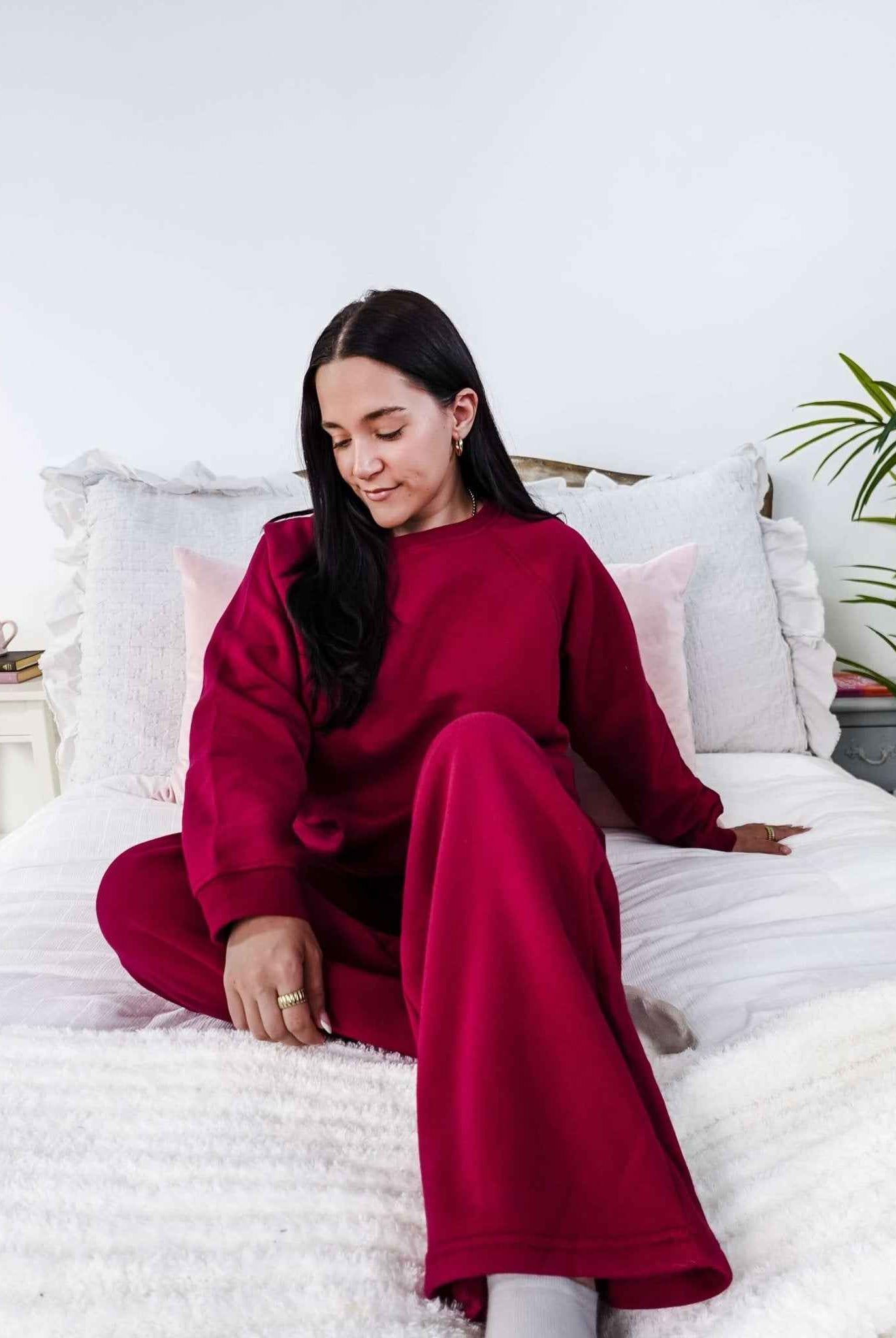 Woman in a red sweatsuit sitting on a bed in a bright room with white walls and a plant.