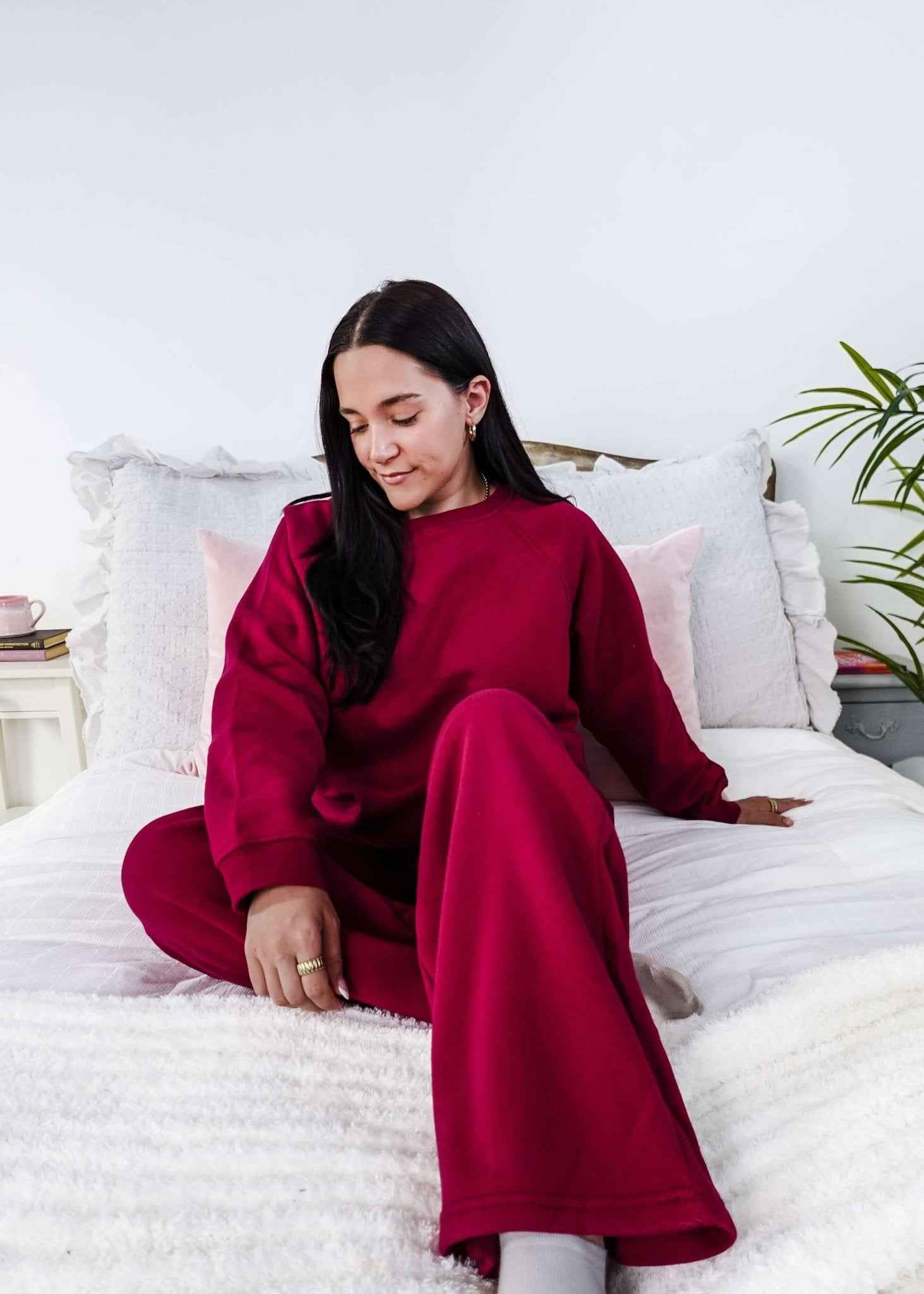 Woman in a red sweatsuit sitting on a bed in a bright room with white walls and a plant.