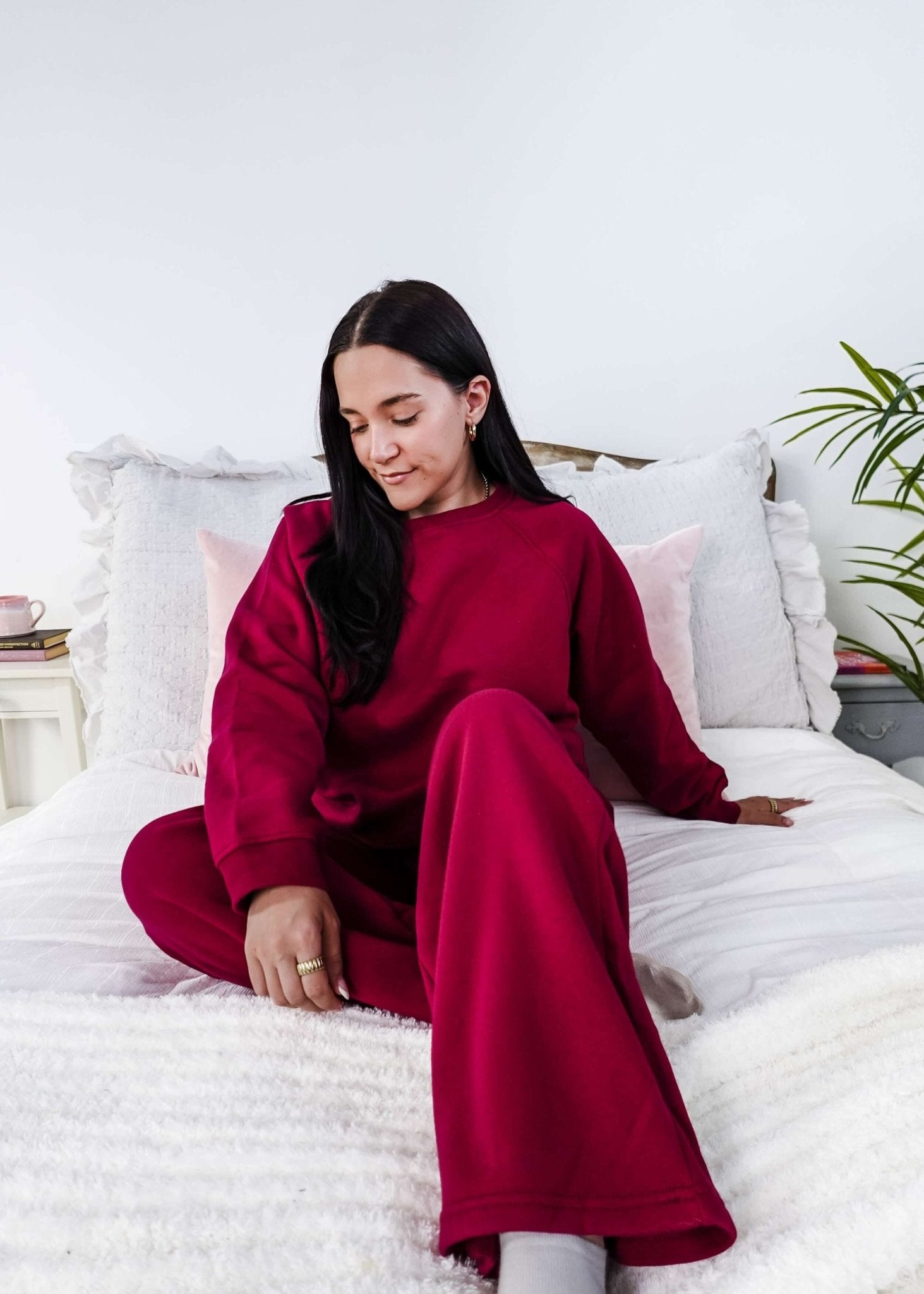 Woman in a red sweatsuit sitting on a bed in a bright room with white walls and a plant.