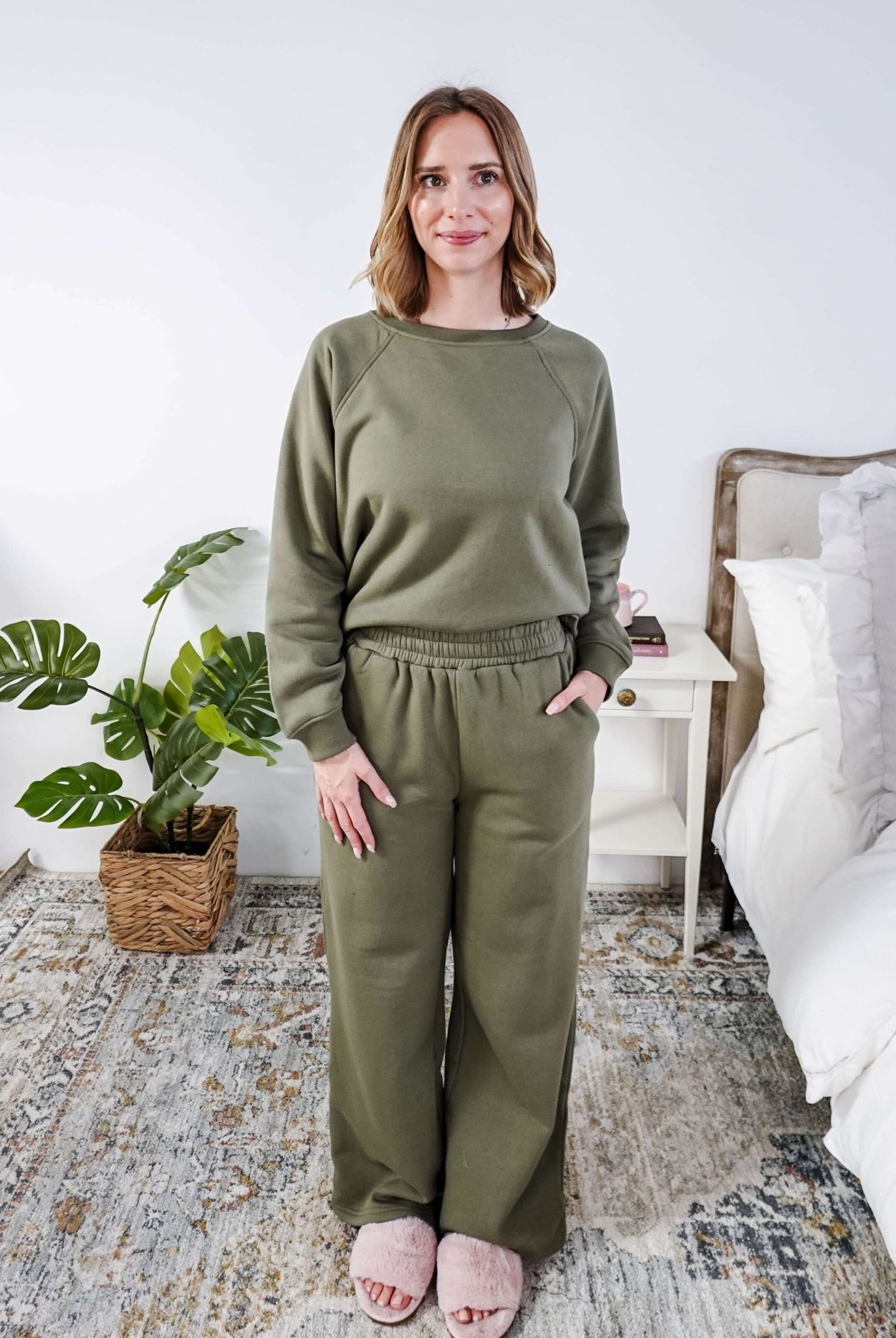 Woman wearing a green sweatsuit outfit standing in a bedroom with a plant and bed in the background.