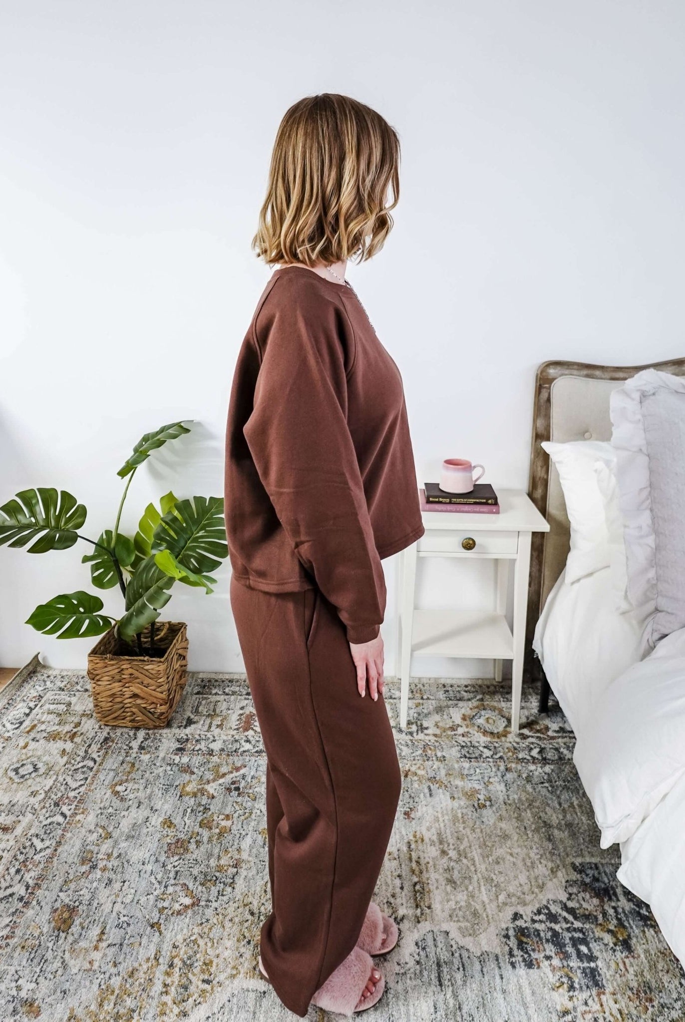 Person wearing a brown lounge outfit standing in a bedroom with a plant and bed in the background.