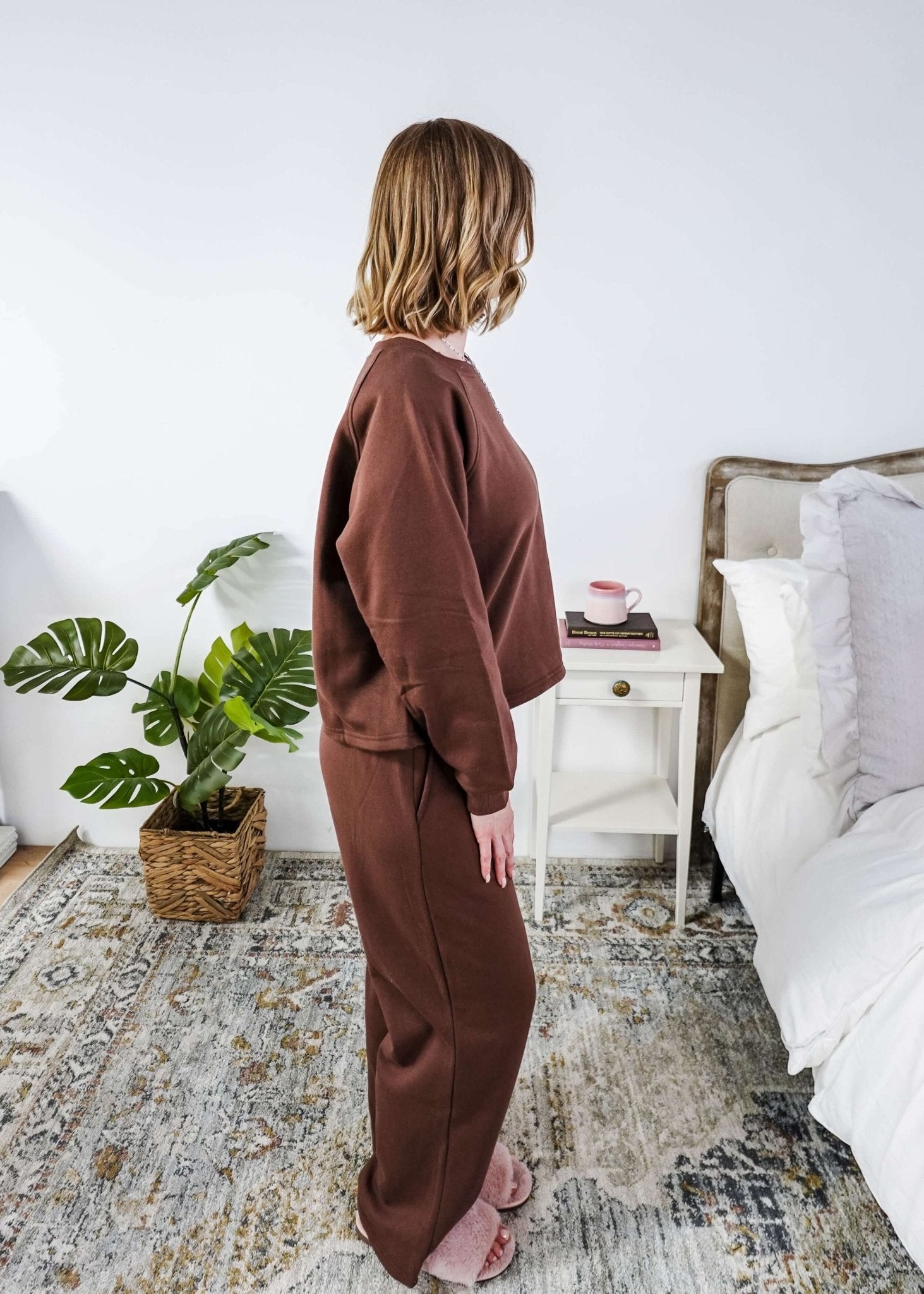 Person wearing a brown lounge outfit standing in a bedroom with a plant and bed in the background.
