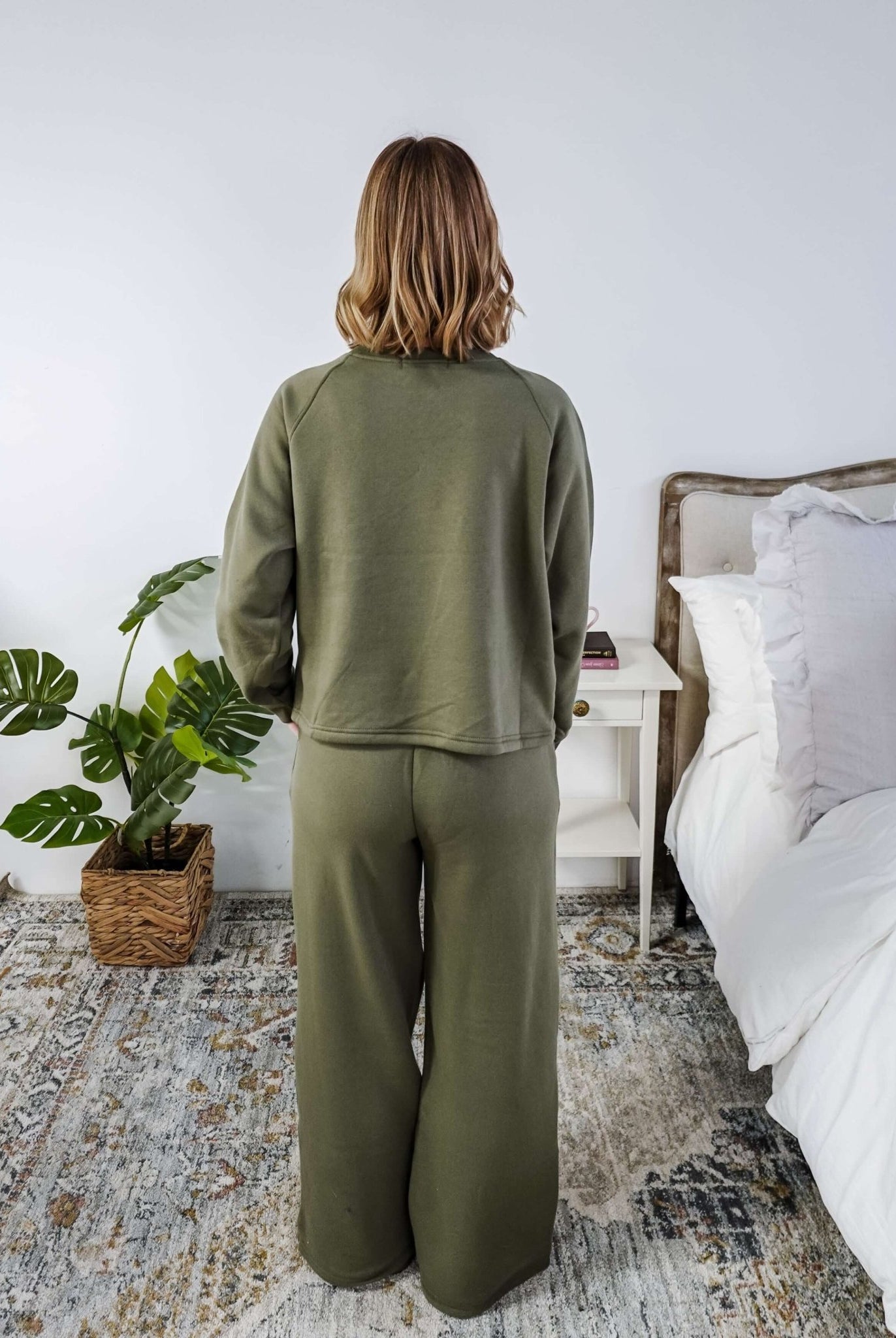 Person wearing a green outfit standing in a bedroom with a bed and plant.