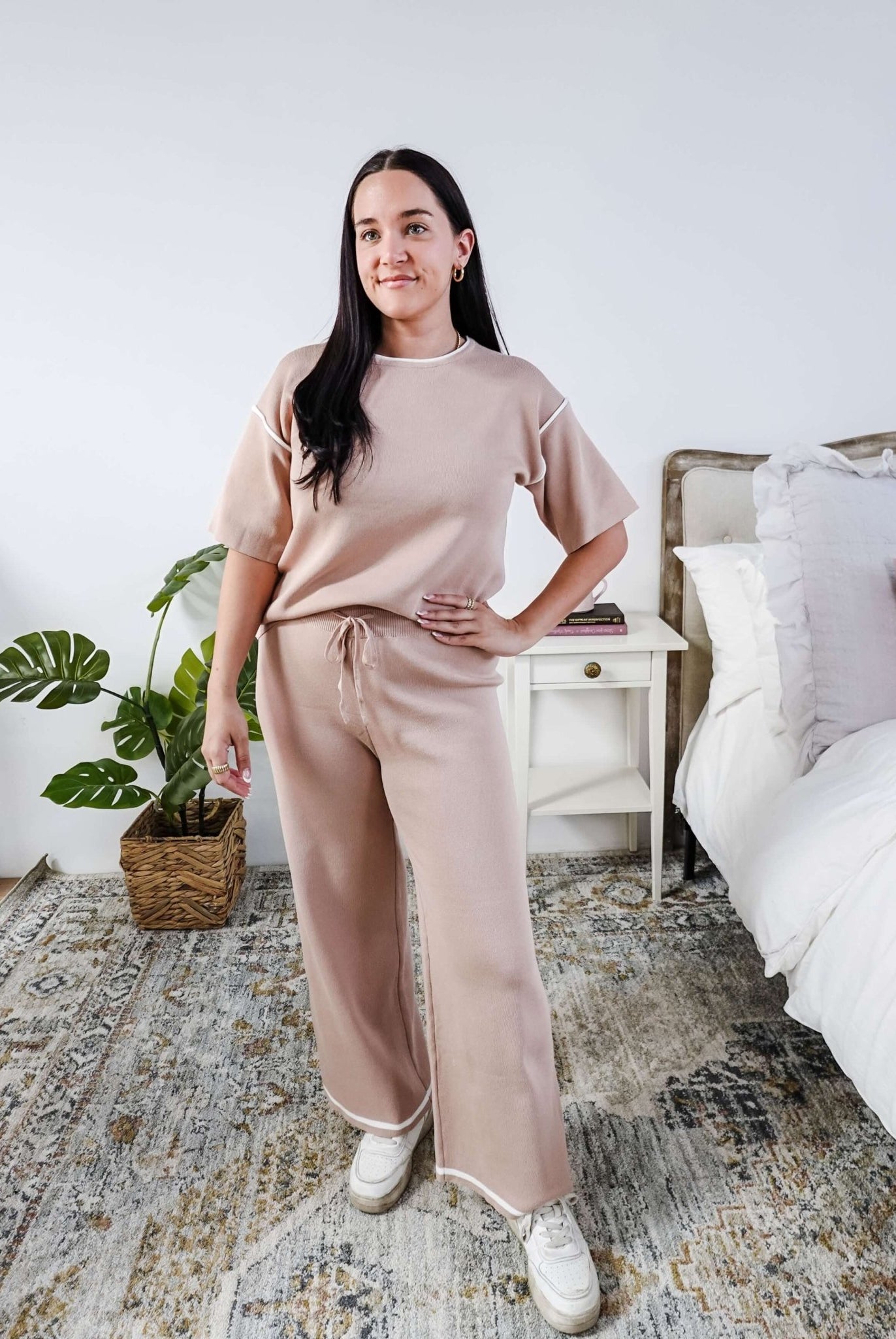 Woman in a beige outfit standing in a bedroom with a plant and bed in the background
