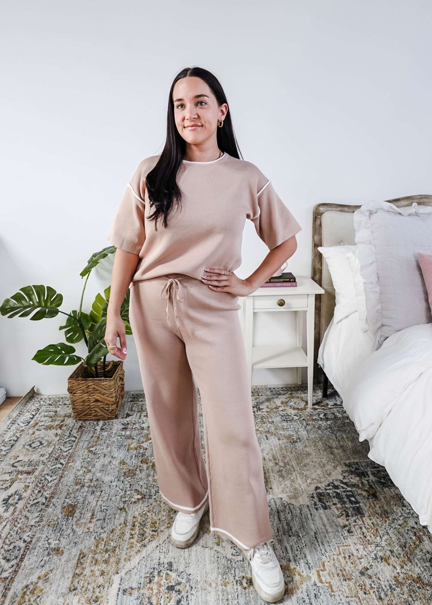 Woman in a beige outfit standing in a bedroom with a plant and bed in the background