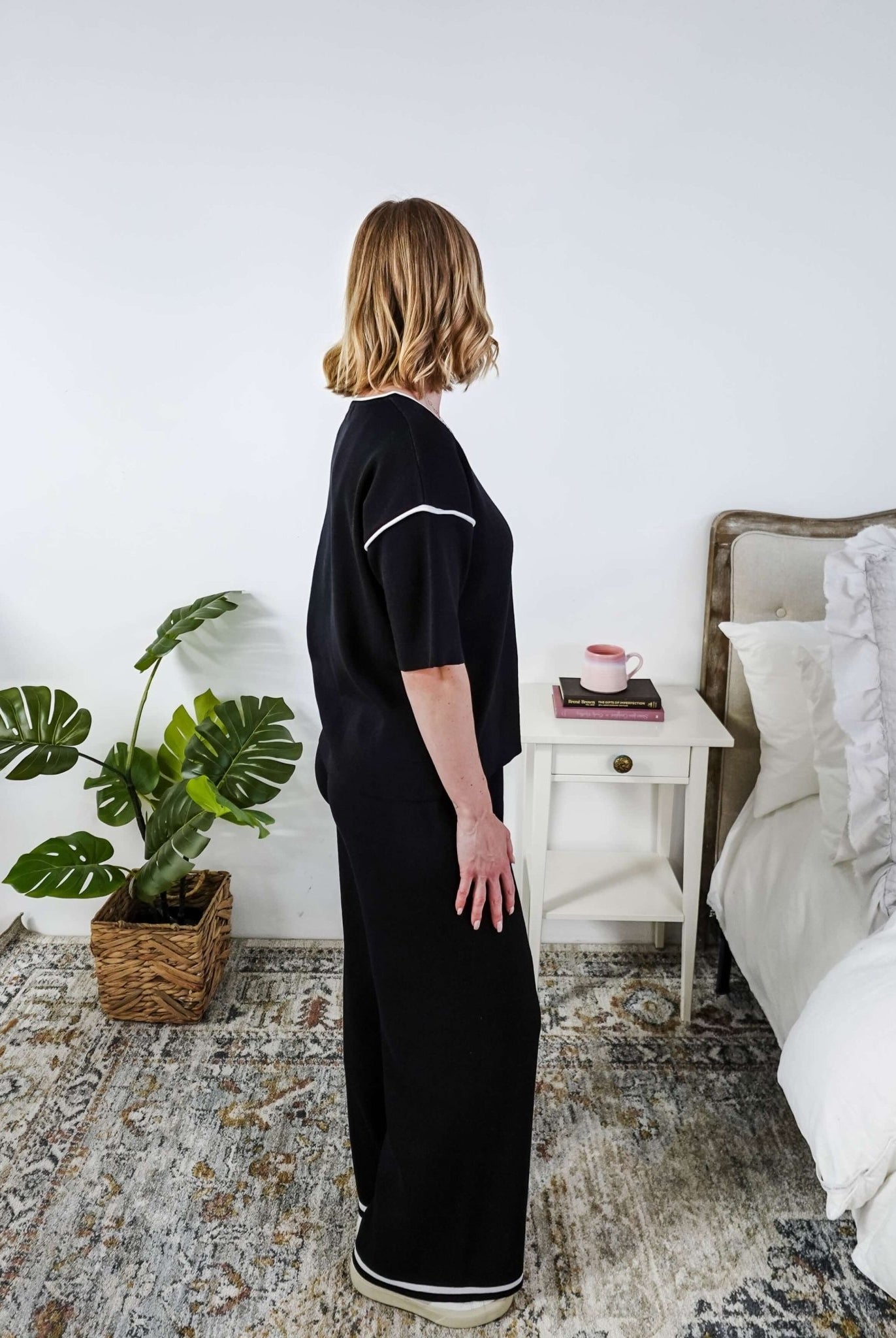 Woman in a black lounge outfit standing in a bedroom with a plant and bed in the background