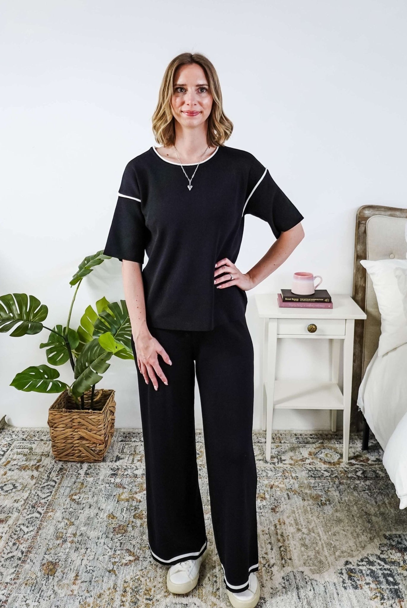 Woman wearing a black outfit in a bedroom setting with a plant and bed in the background.
