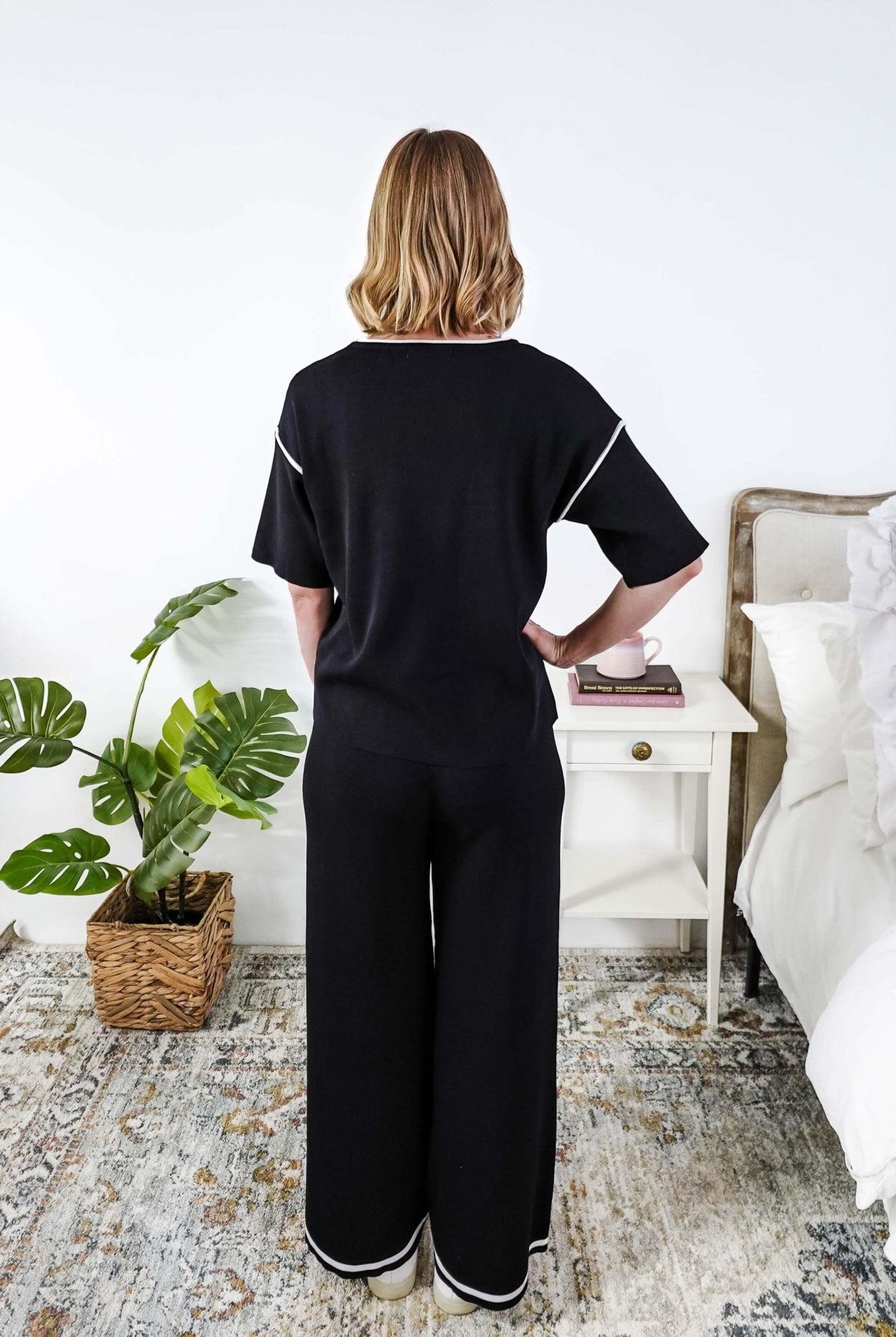 Person wearing a black lounge outfit standing in a bedroom with a plant and nightstand.