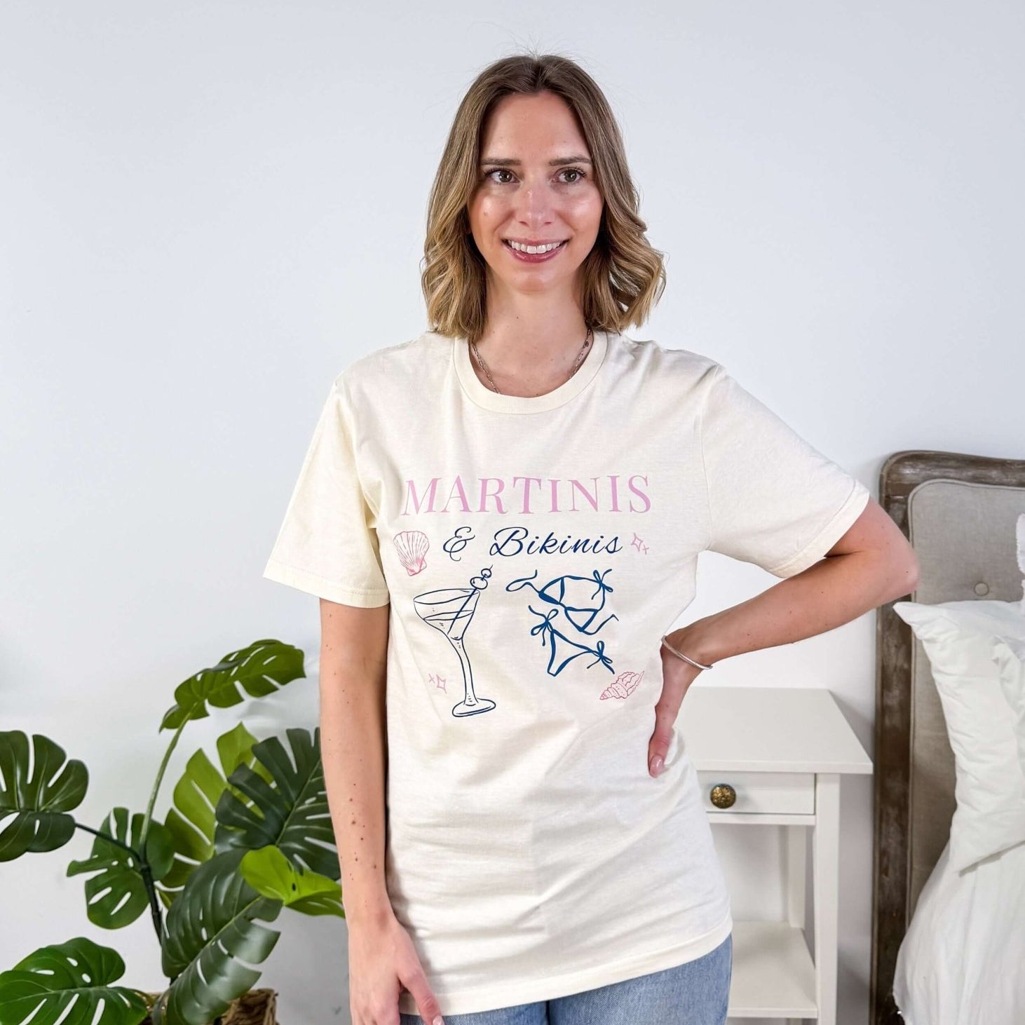 Martinis & Bikinis Relaxed Fit Graphic Tee with martini and bikini print in pink and blue and some seashells, worn on model with jeans in a bedroom setting