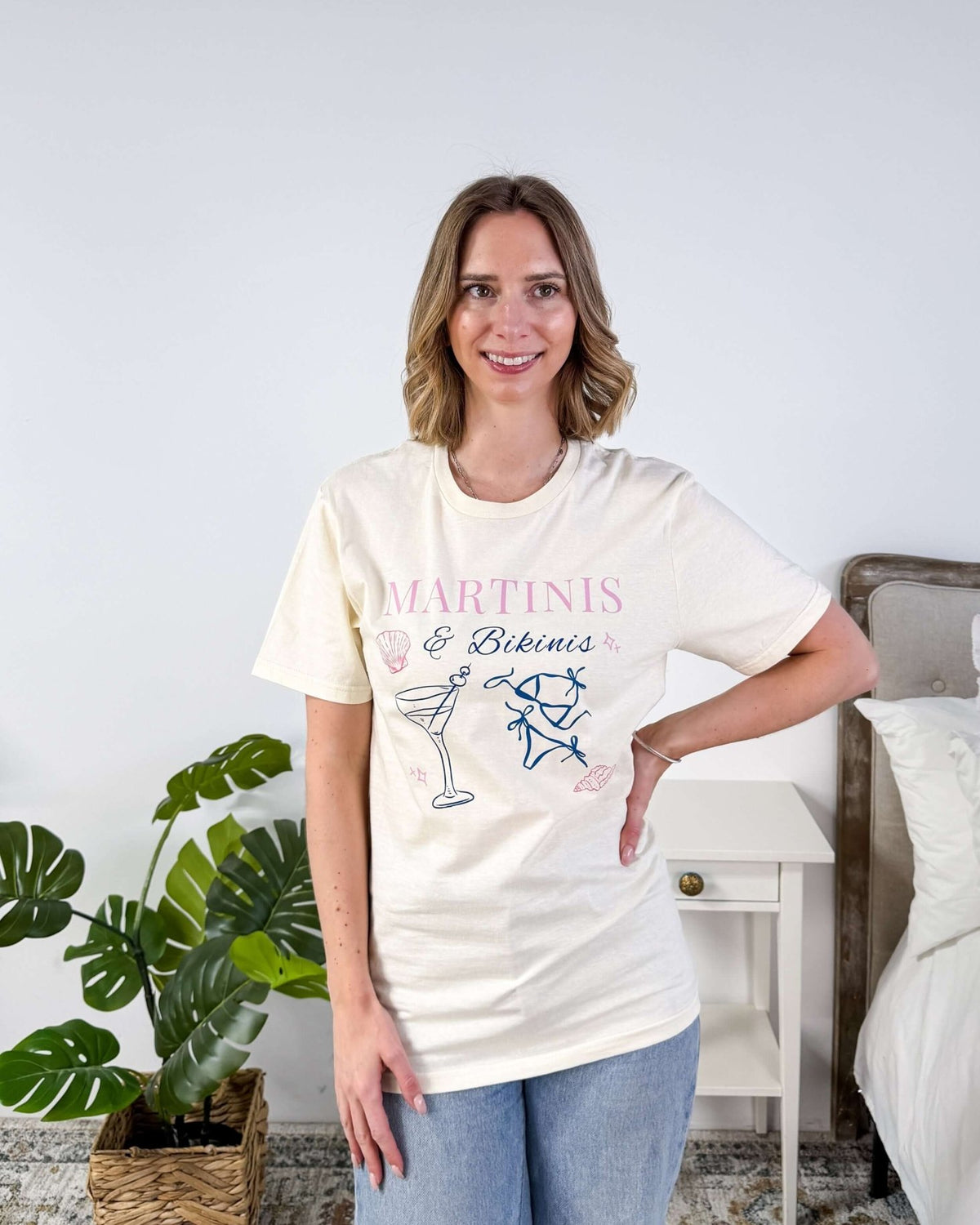 Martinis & Bikinis Relaxed Fit Graphic Tee with martini and bikini print in pink and blue and some seashells, worn on model with jeans in a bedroom setting