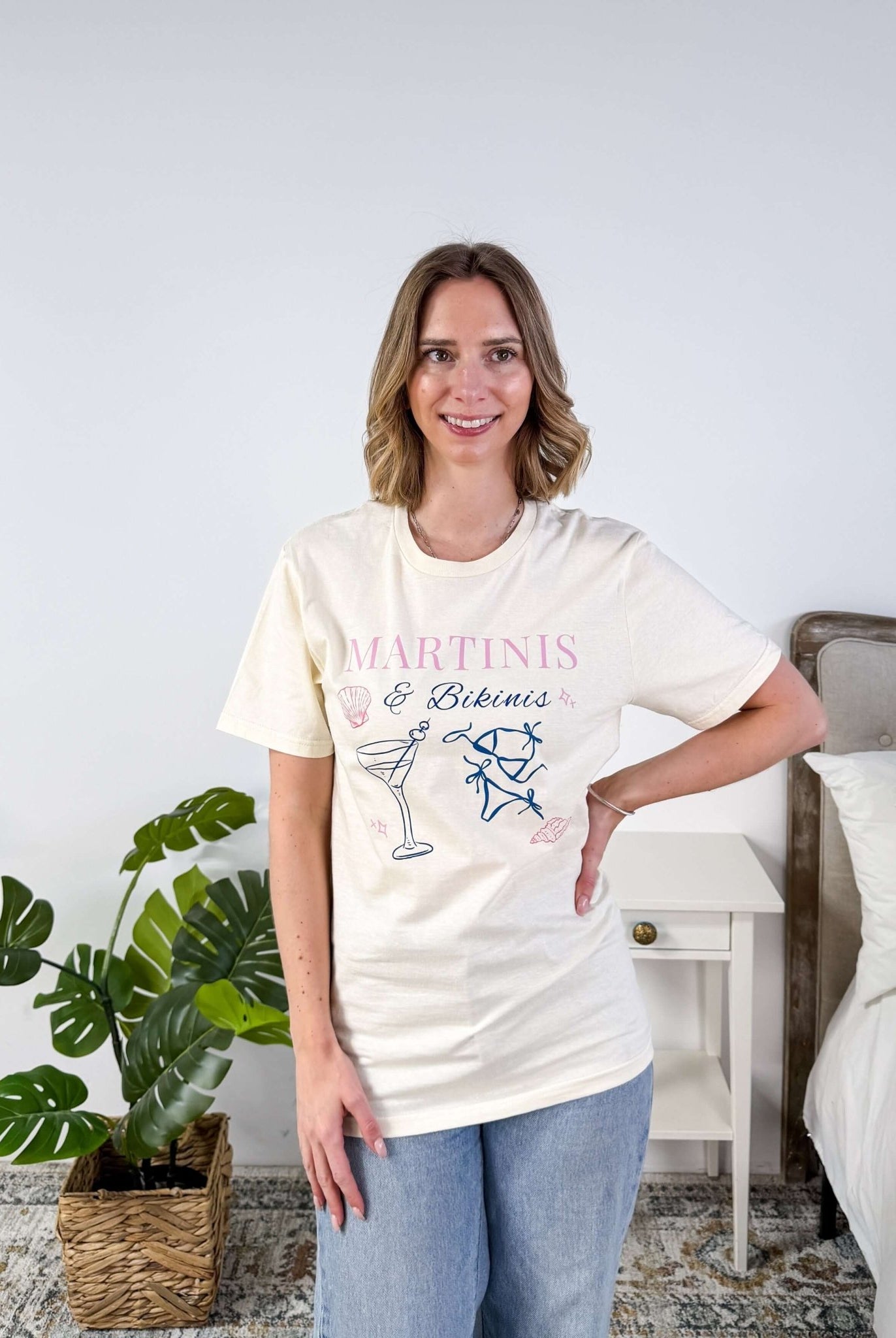 Martinis & Bikinis Relaxed Fit Graphic Tee with martini and bikini print in pink and blue and some seashells, worn on model with jeans in a bedroom setting