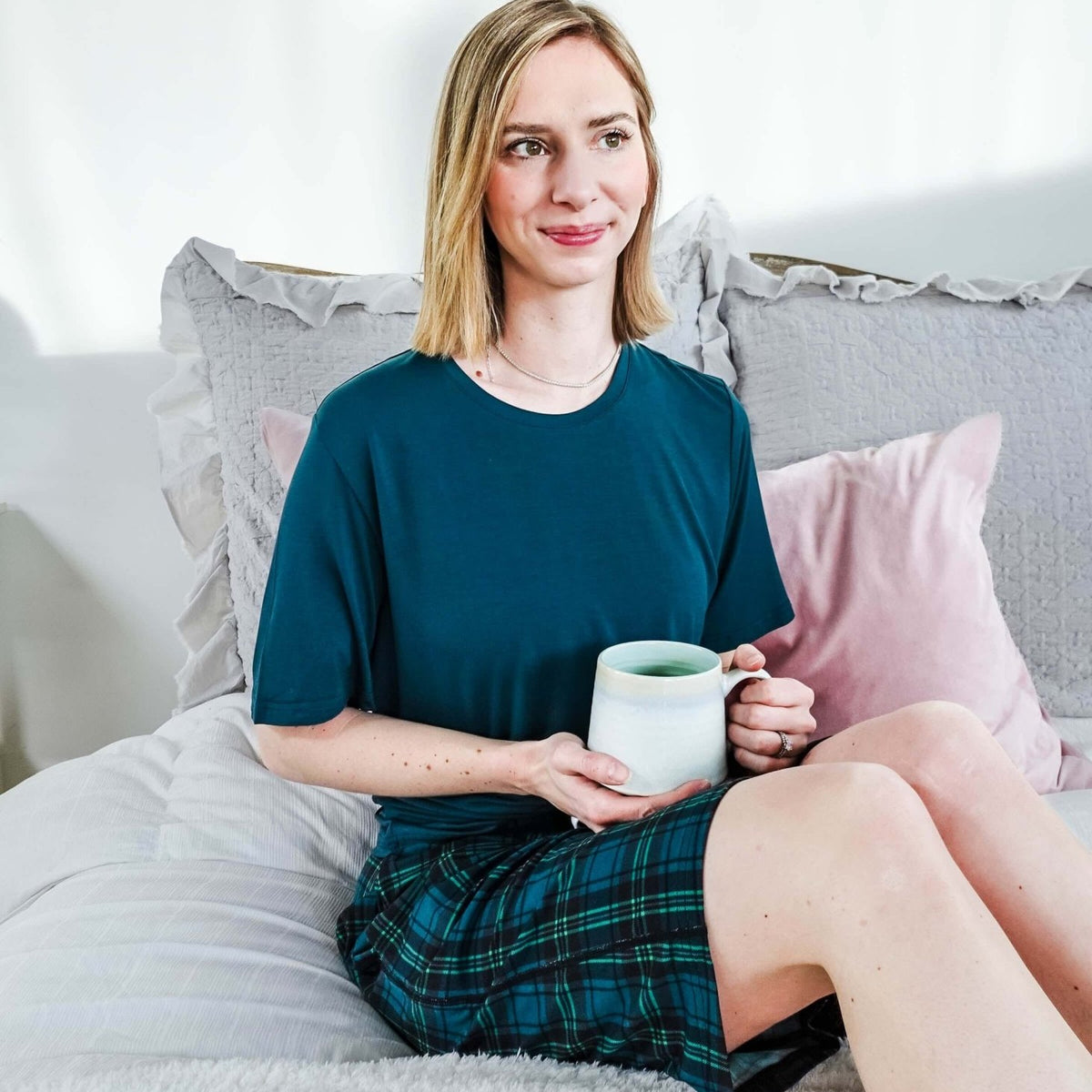 A woman sitting on a bed in Midnight Plaid Unisex Bamboo Sleep Shorts, holding a mug, enjoying a cozy atmosphere.