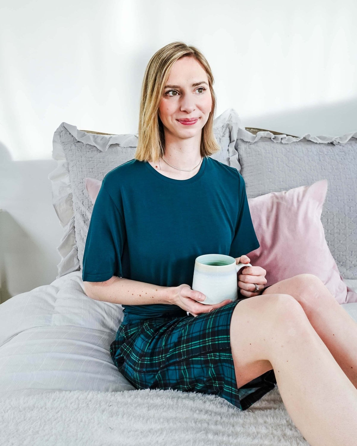A woman sitting on a bed in Midnight Plaid Unisex Bamboo Sleep Shorts, holding a mug, enjoying a cozy atmosphere.