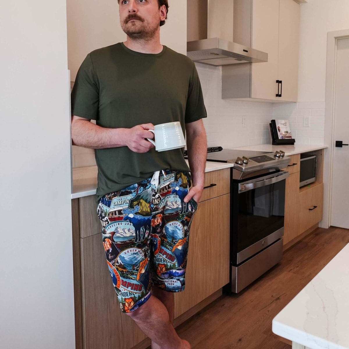 Man standing in kitchen wearing Mountains Are Calling Print Unisex Shorts Bamboo and a green t-shirt, holding a coffee cup.