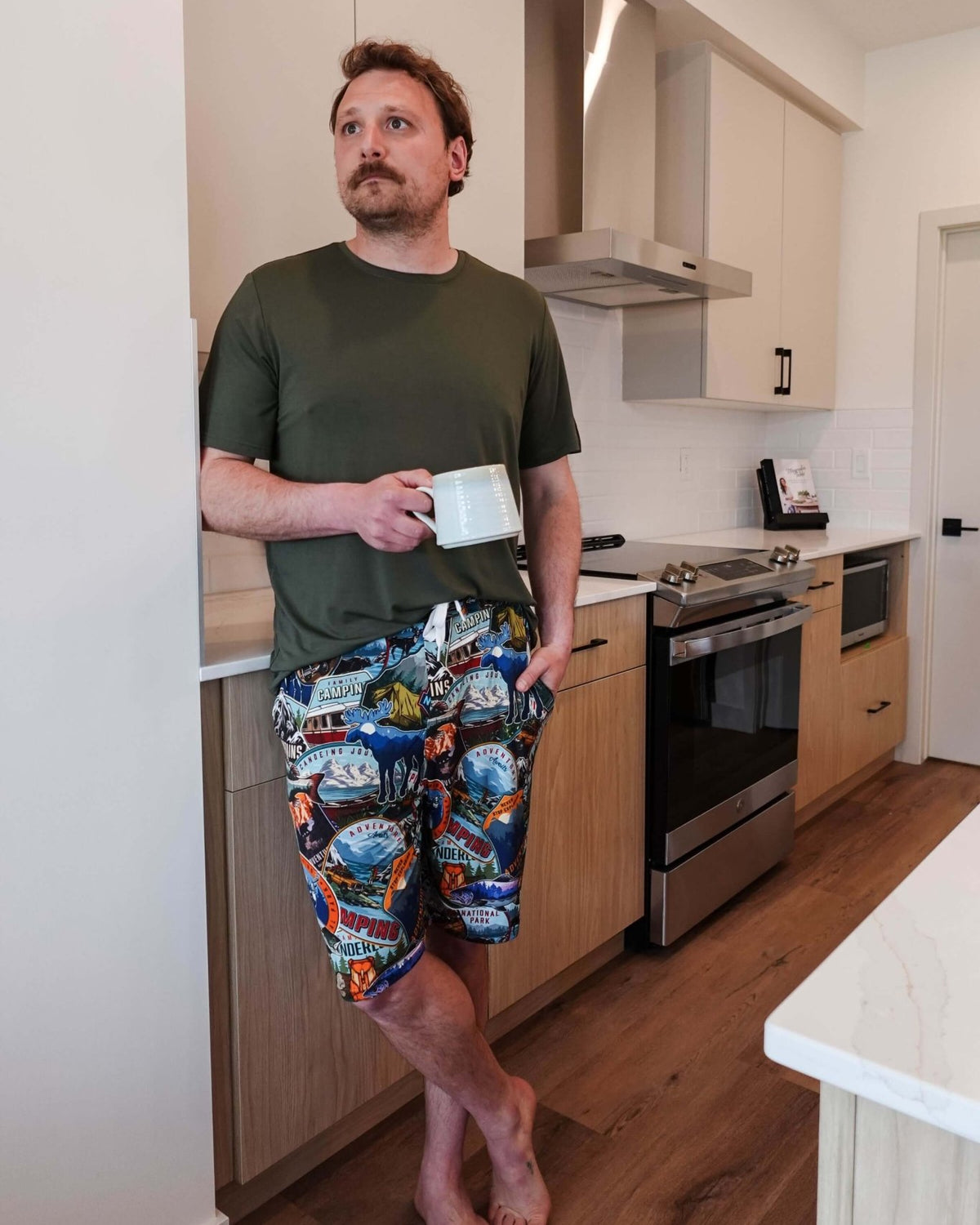Man standing in kitchen wearing Mountains Are Calling Print Unisex Shorts Bamboo and a green t-shirt, holding a coffee cup.
