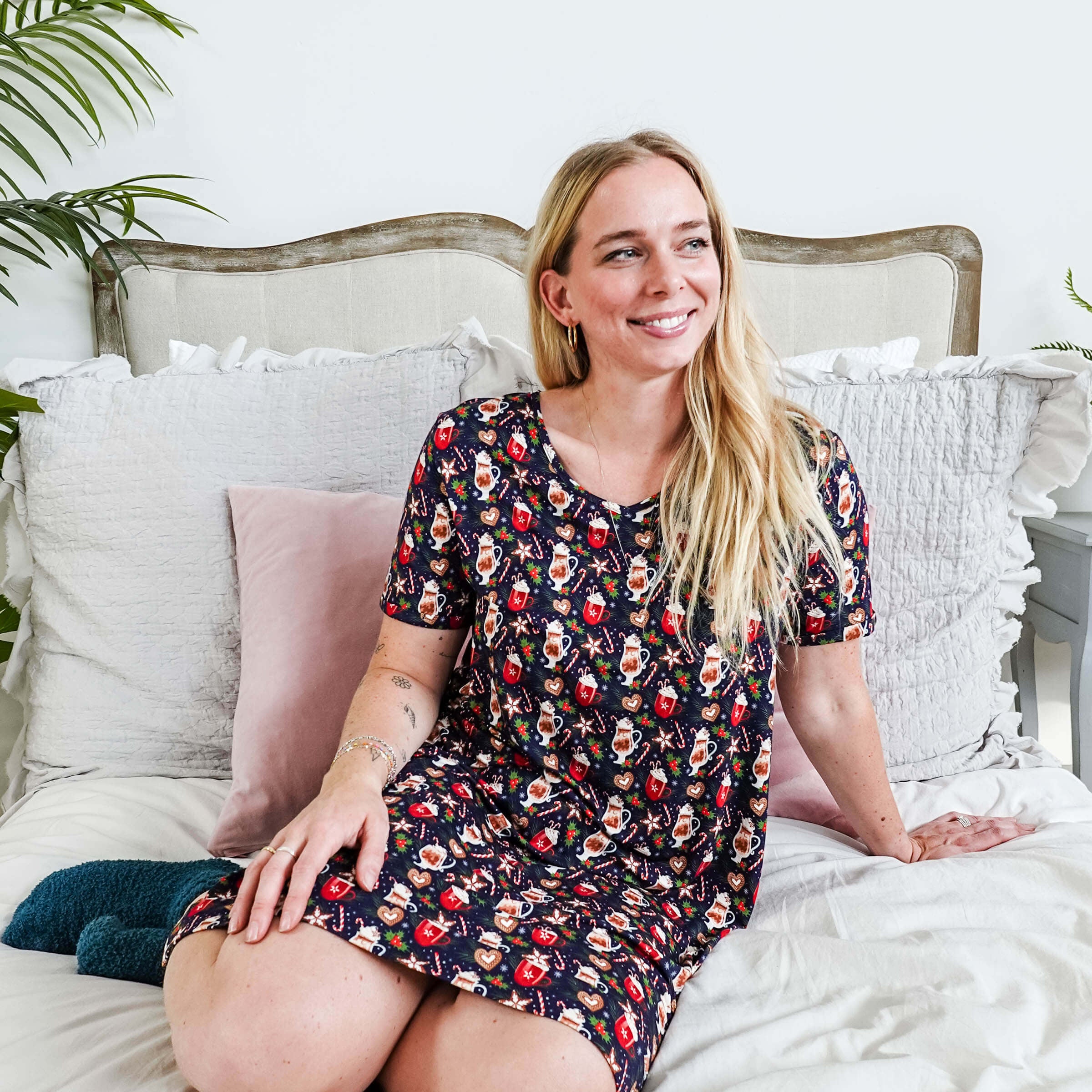 Woman sitting on bed wearing Merry Mugs V-Neck Bamboo Nightshirt with a holiday print and pockets by Georgia Rylan.