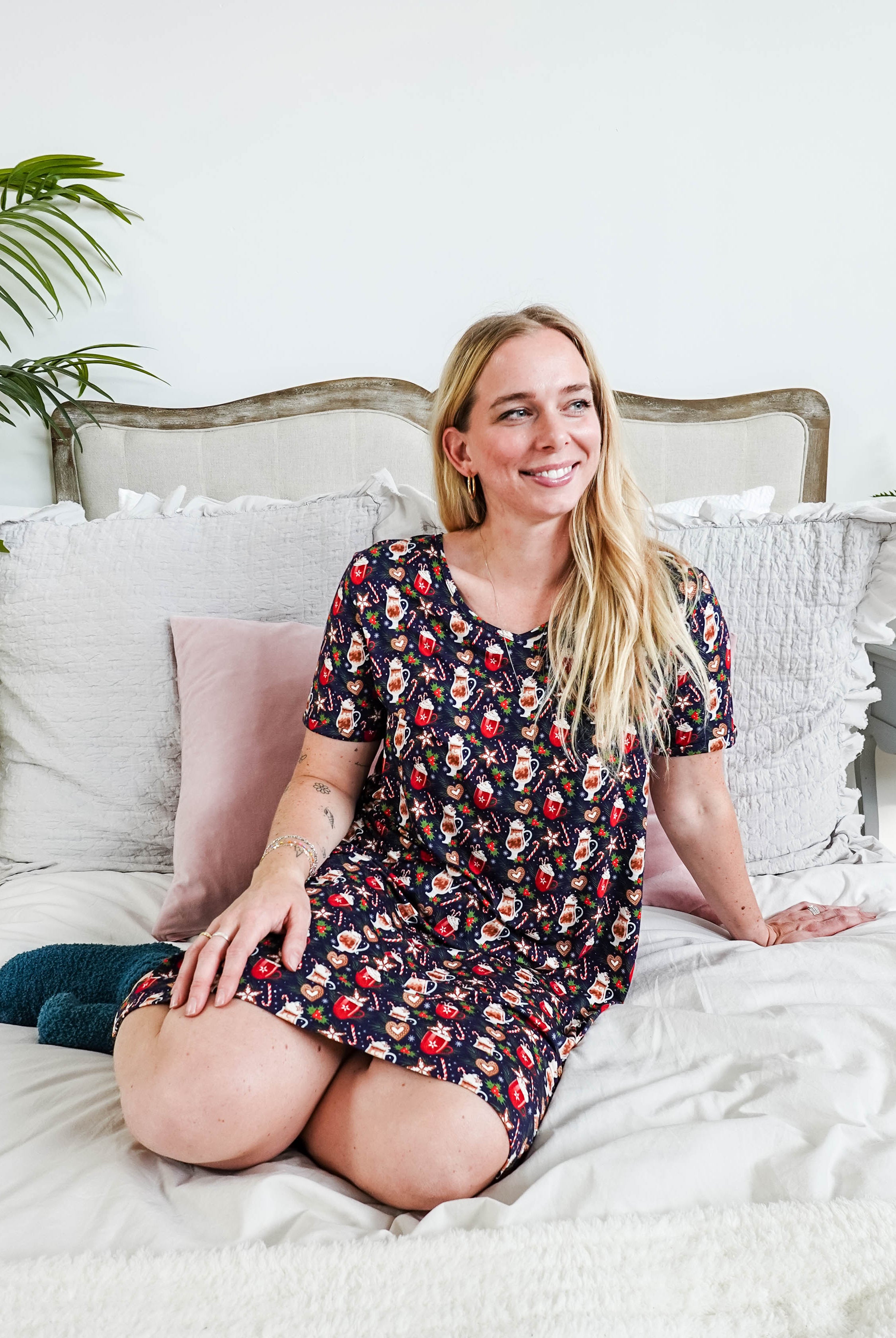Woman sitting on bed wearing Merry Mugs V-Neck Bamboo Nightshirt with a holiday print and pockets by Georgia Rylan.