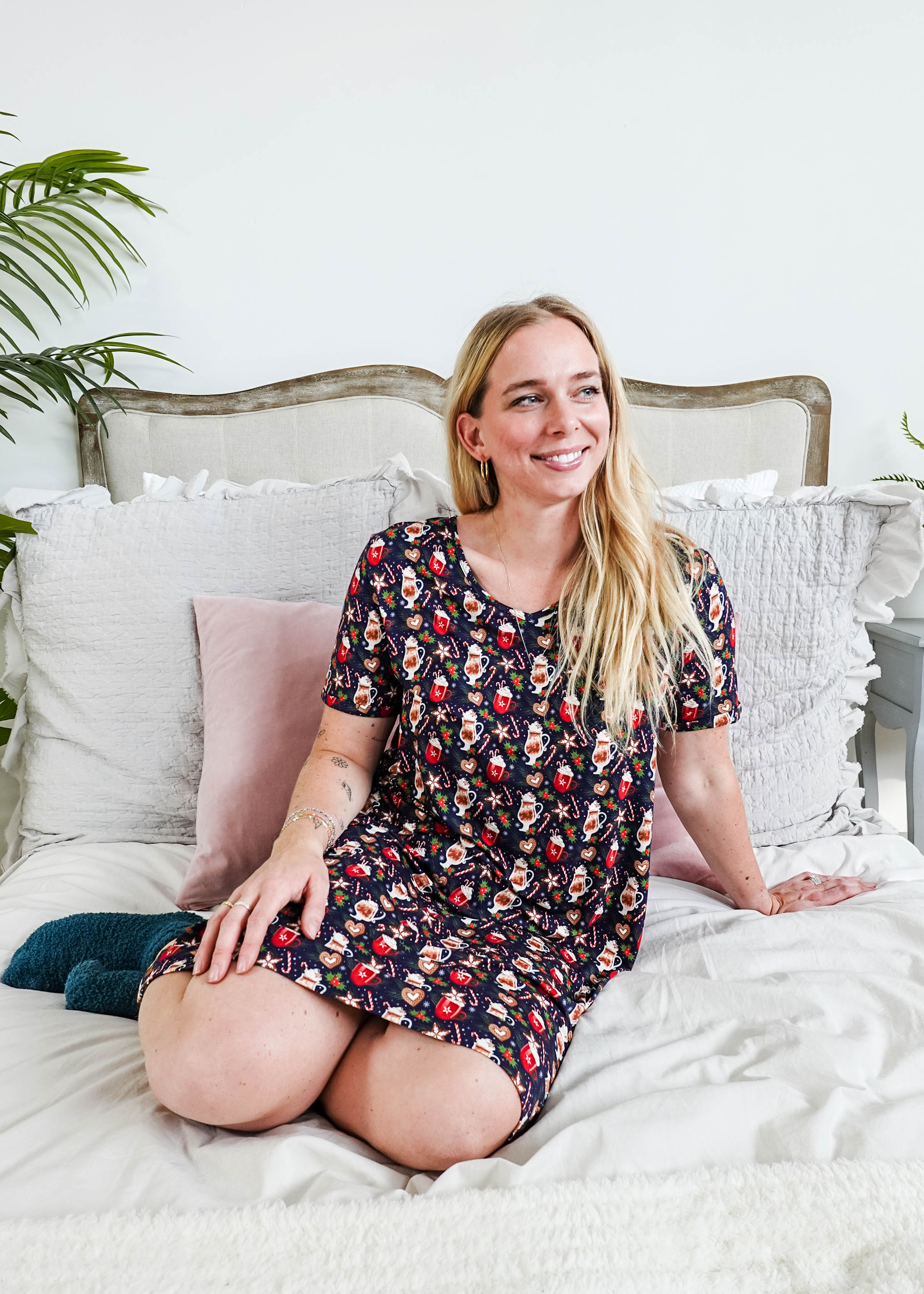 Woman sitting on bed wearing Merry Mugs V-Neck Bamboo Nightshirt with a holiday print and pockets by Georgia Rylan.