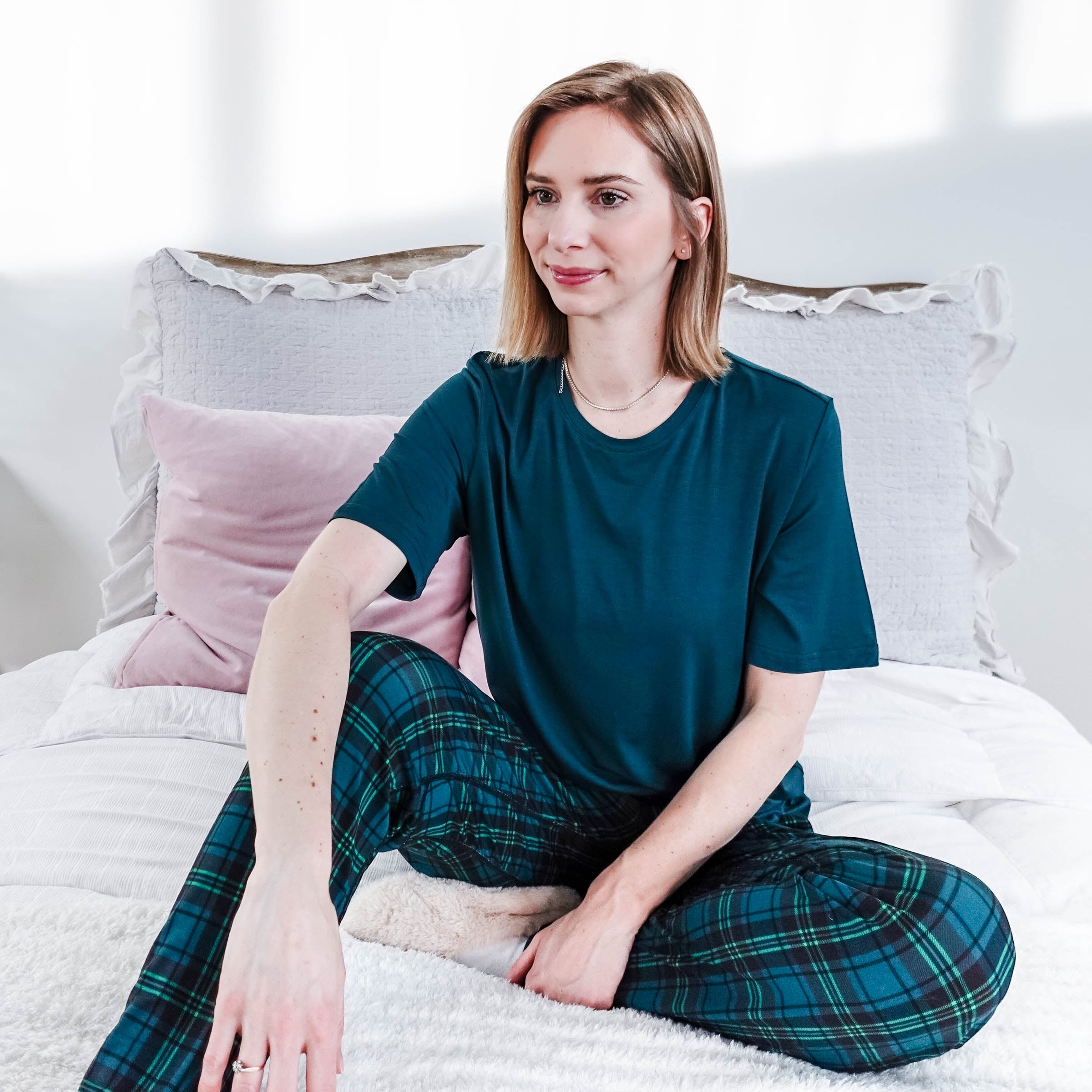 Model wearing Midnight Plaid Unisex Bamboo Joggers and a teal shirt, sitting on a bed with soft pillows.
