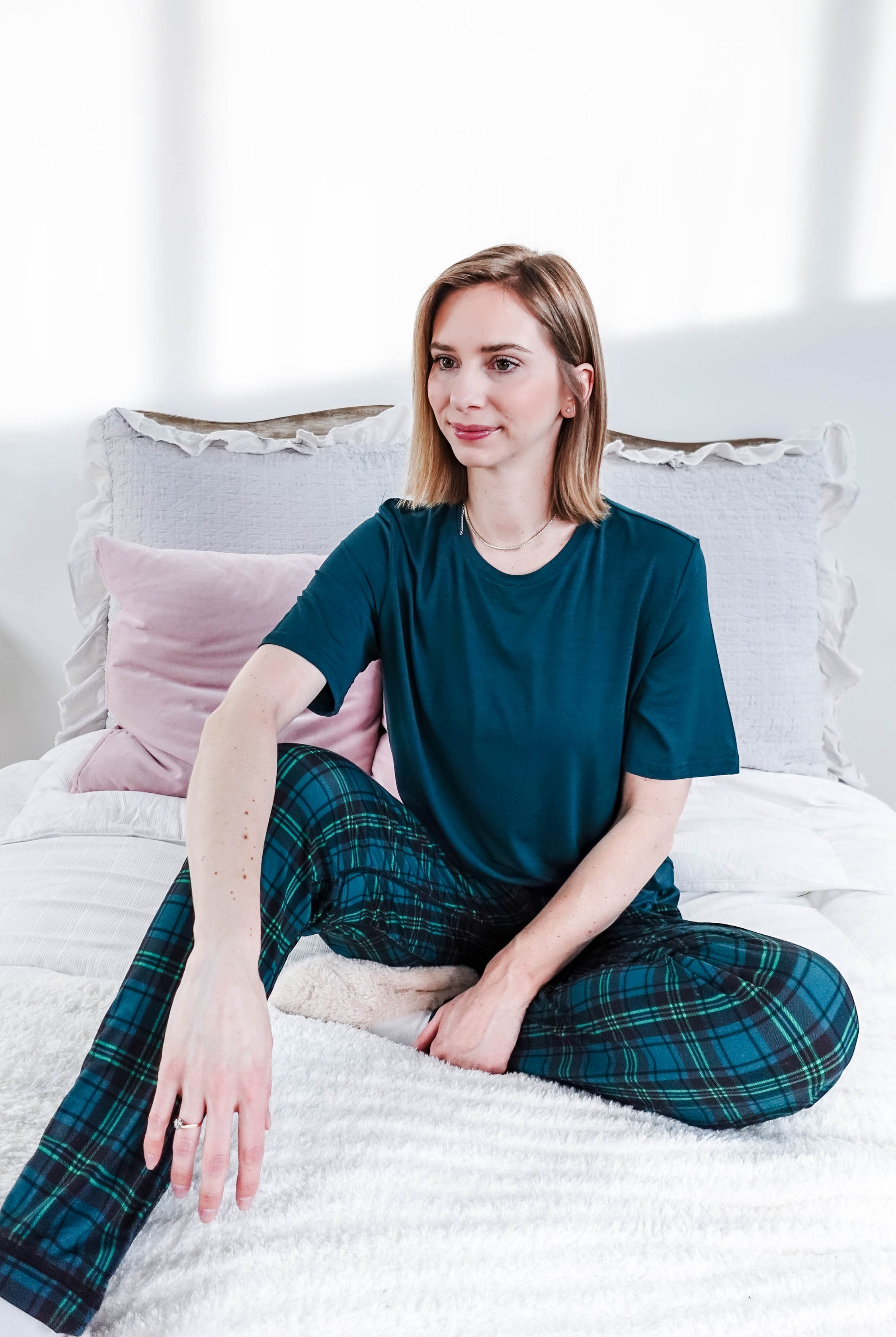 Model wearing Midnight Plaid Unisex Bamboo Joggers and a teal shirt, sitting on a bed with soft pillows.