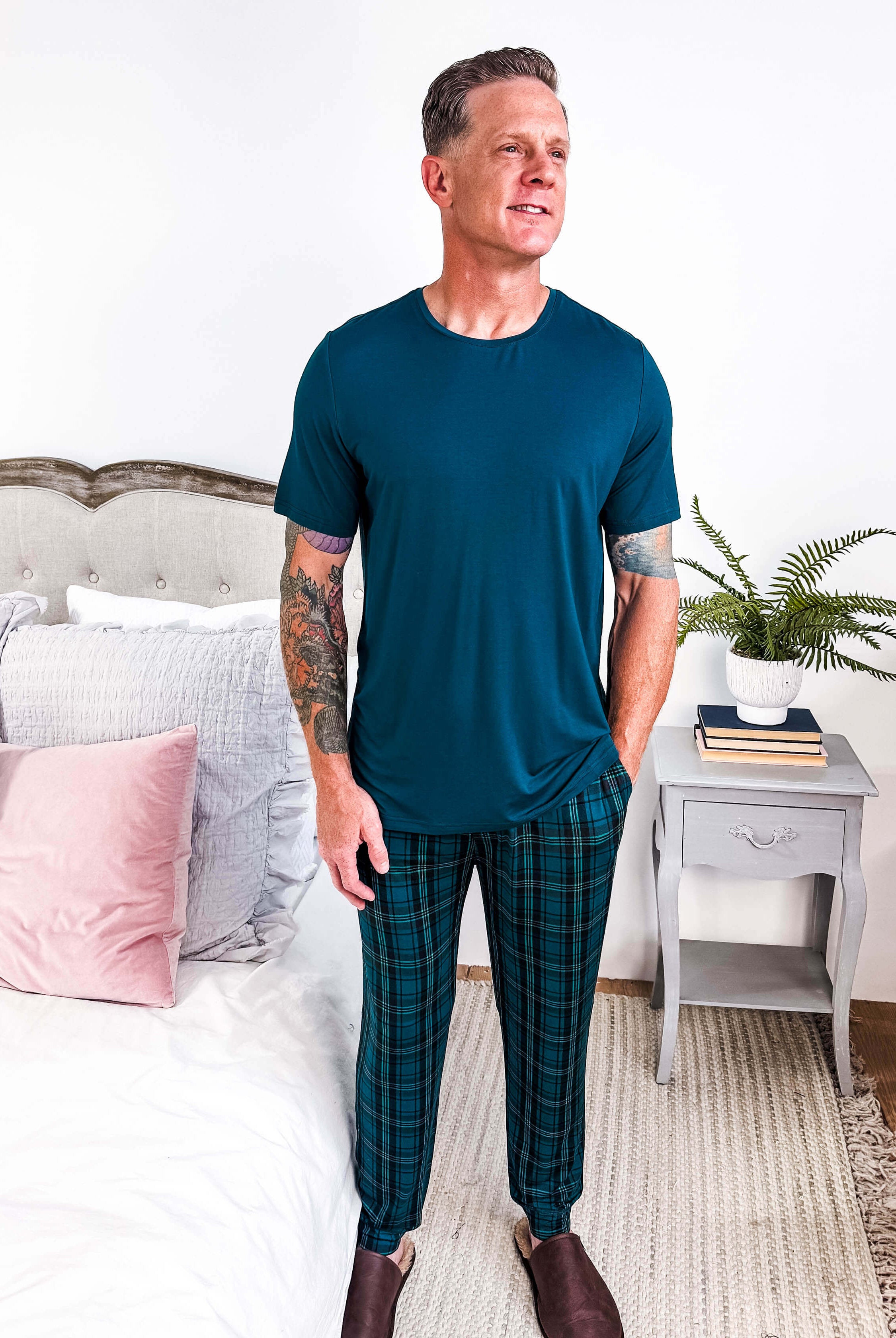 Model wearing Midnight Plaid Unisex Bamboo Joggers paired with a blue shirt in a cozy bedroom setting.