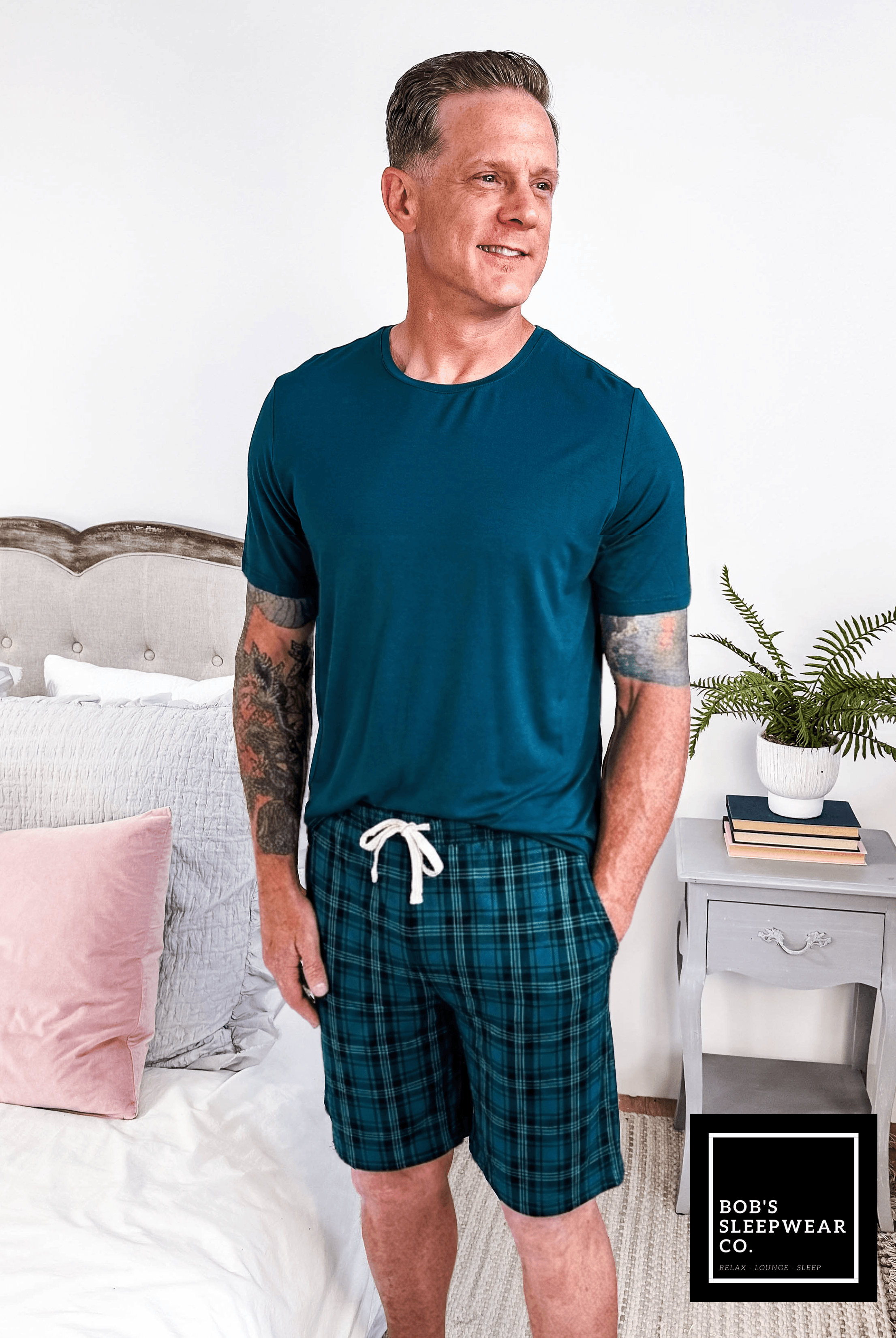 Man wearing Midnight Plaid Unisex Bamboo Sleep Shorts and a blue T-shirt in a cozy bedroom setting.
